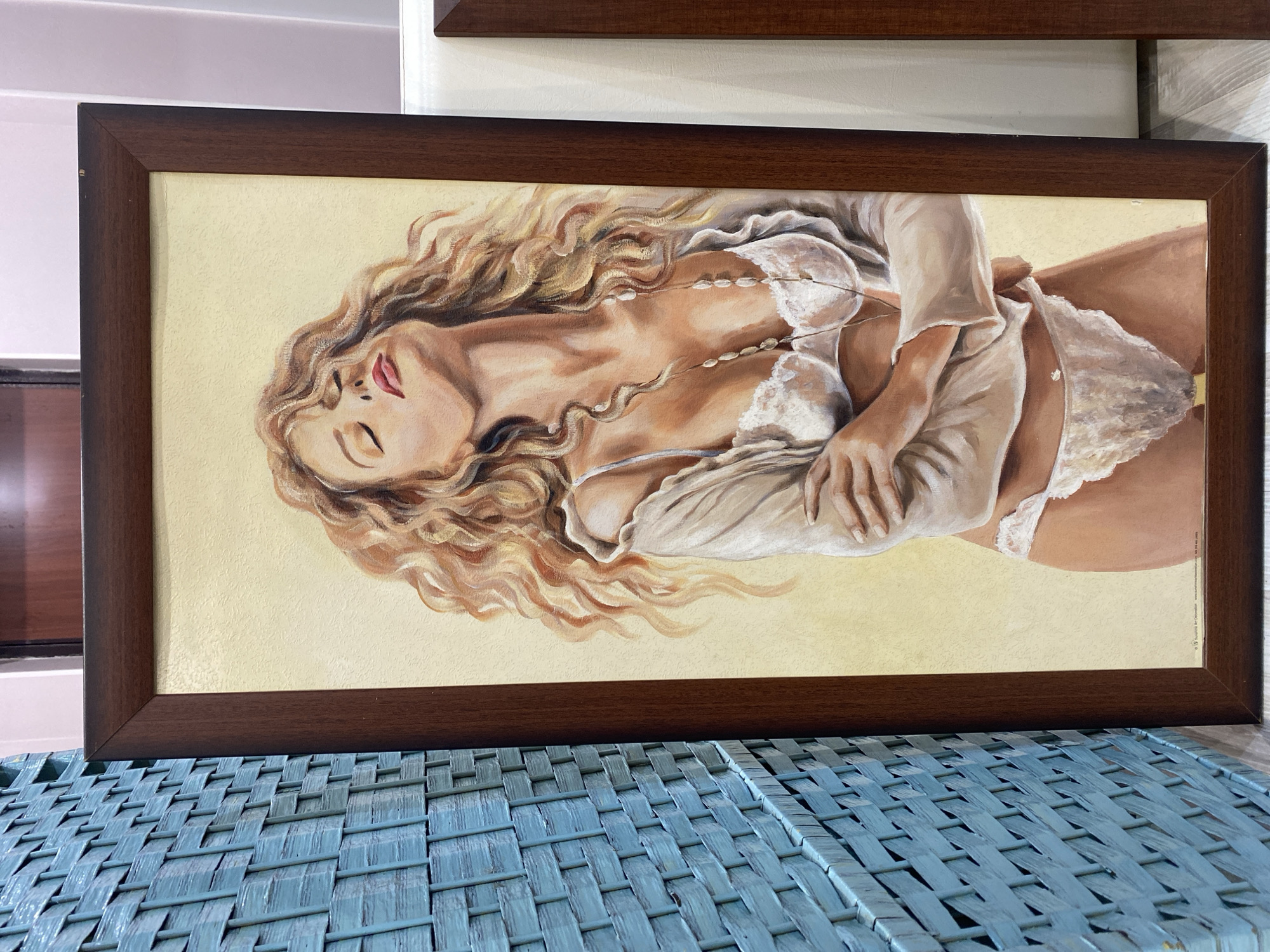 A framed painting of a reclining woman with wavy blonde hair is positioned on top of a woven blue surface. The woman is wearing a white garment and lingerie. The painting has a dark brown frame. The painting is indoors.