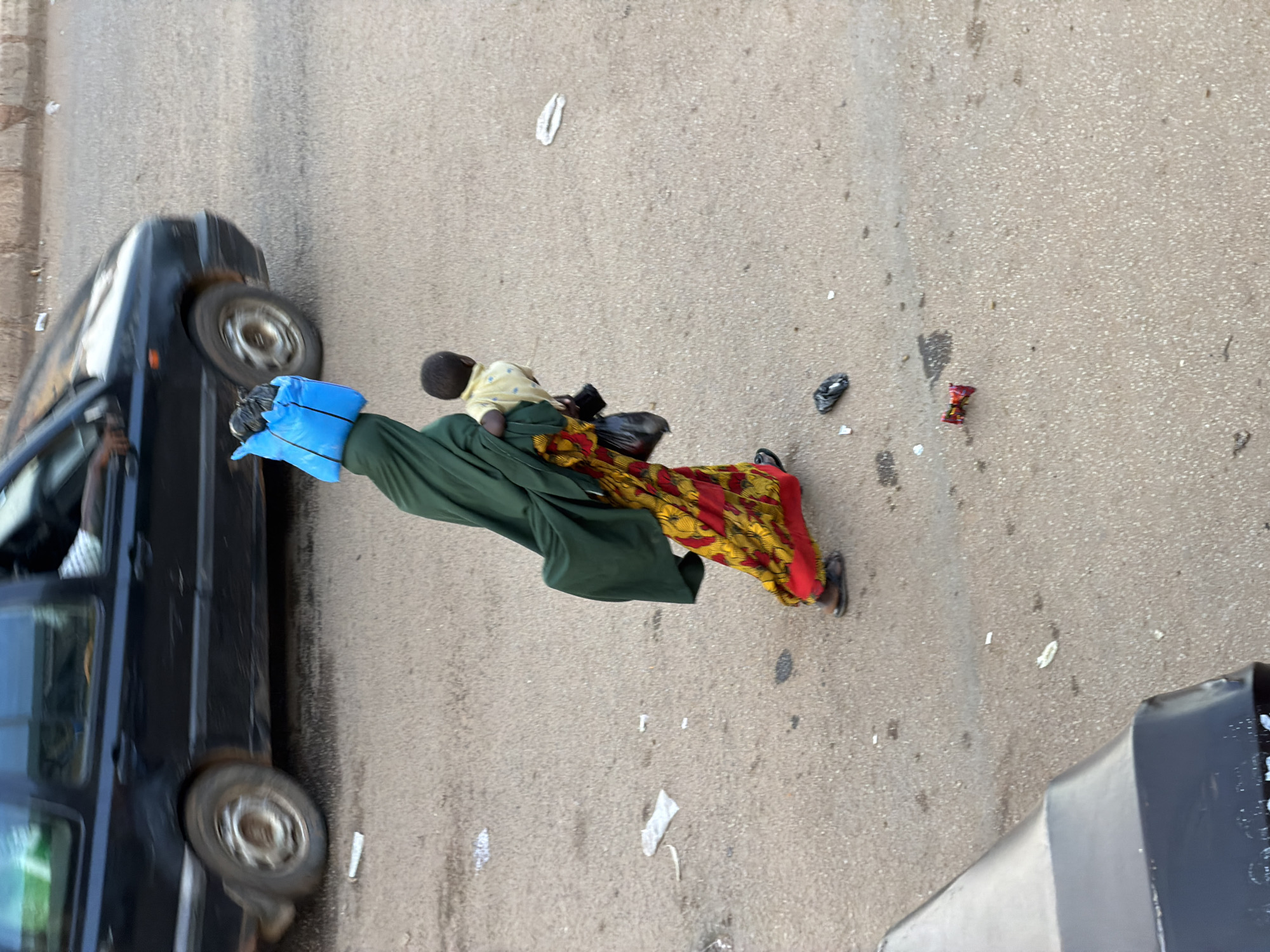 This media file shows a scene on a paved road in Bukuru, Nigeria. The main subjects are two people, an adult and a child, and a black car. The adult is carrying the child, who is wearing a light yellow shirt. The adult is draped in a long green garment, with a colorful patterned fabric (red, yellow, and black) visible around their legs. They are also wearing dark sandals. The black car appears to be a van or SUV and is parked or moving slowly on the left side of the frame. The road surface is asphalt and shows signs of wear, with scattered debris, including plastic wrappers and what appears to be a red candy wrapper.
The image is taken from a high angle, possibly from a window or another vehicle. The lighting suggests it is daytime, with natural light illuminating the scene. There are no clear indicators of the weather, but the light suggests it is not overcast.
The adult appears to be holding the child securely, with the child resting against their chest. The overall impression is a moment of transit or a brief stop on the roadside. The presence of debris suggests a public area where litter is common. The context of Bukuru, Nigeria, is provided as a location cue. No text is visible in the image.