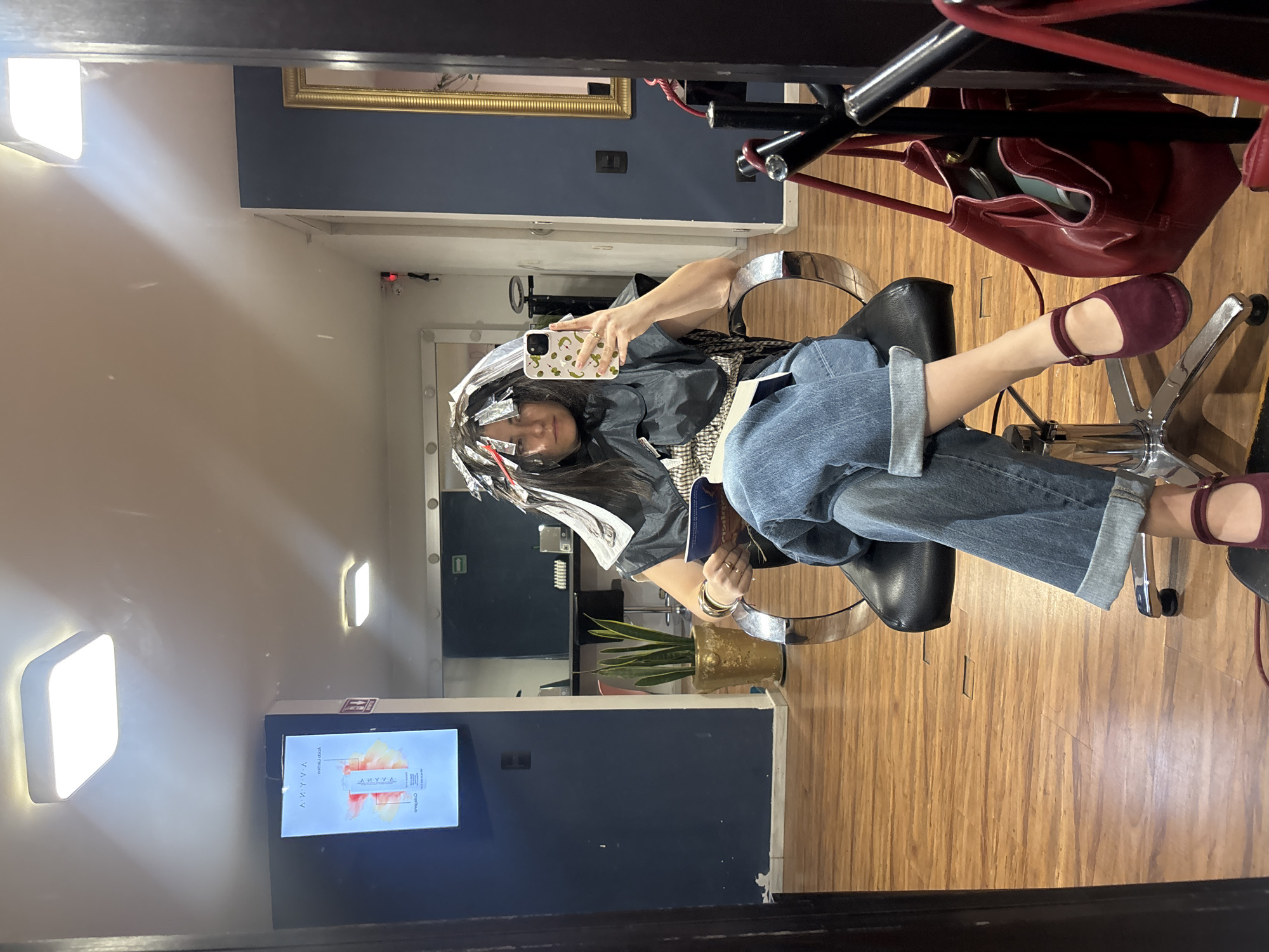 The image shows a woman taking a mirror selfie in a hair salon. She is seated in a black salon chair, wearing a black protective cape over a grey top, blue cuffed jeans, and burgundy flat shoes. Her dark hair is sectioned with numerous silver foils, indicating she is undergoing a hair coloring or highlighting treatment. She holds a smartphone with a light-colored case adorned with a pattern of small peppers or similar designs.
The setting is an indoor space with light wooden flooring. The walls are painted in two tones: a dark blue accent wall and a lighter cream or white. A large, ornate golden-framed mirror hangs on the dark blue wall. To the woman's left, a snake plant sits in a golden pot. A red handbag hangs on the back of her chair. On a wall to her right, a digital screen displays an advertisement for a beauty product, featuring a bottle and the mirrored text "ANAYYA" (likely "AYYANA" when un-mirrored) and another word that appears to be "Serum." The woman looks relaxed, with her legs crossed, and she appears to be holding a book or magazine in her lap. The lighting is bright, provided by wall-mounted fixtures. The overall scene suggests a typical visit to a professional hair salon in Puebla, Mexico.