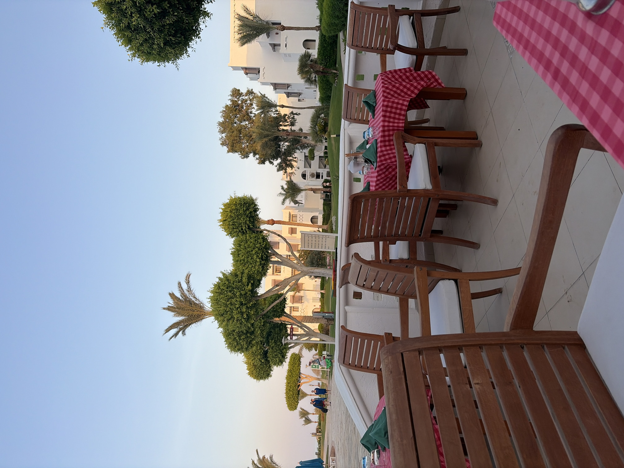 The image depicts an outdoor dining area at a resort in Hurghada, Egypt, during daylight. In the foreground, several wooden chairs with white cushions surround tables covered with red and white checkered tablecloths. Two tables are set with green napkins and glassware. In the midground, a white building with multiple stories and arched windows is visible, surrounded by well-maintained greenery and palm trees. Several people are walking in the background on a paved pathway. The sky is a clear, pale blue, indicating fair weather.