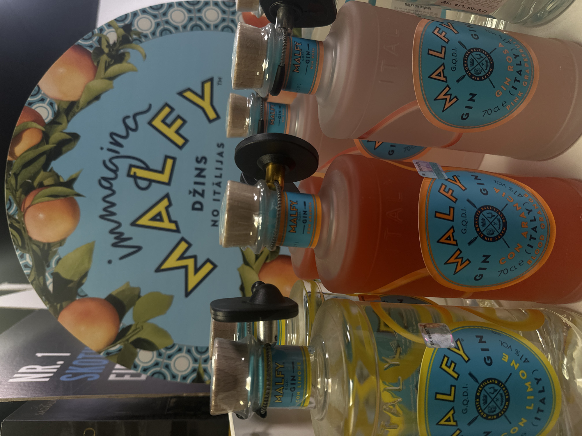The image, rotated 90 degrees clockwise, captures a retail display of Malfy gin bottles. No people are visible, and the scene appears to be an indoor commercial space, likely a store or promotional event in Riga, Latvia, indicated by the visible Latvian text.
The central focus is a stack of at least four Malfy gin bottles, with three distinct flavors clearly identifiable. From top to bottom in the rotated image, these include a frosted bottle of Malfy Gin Rosa (Pink Grapefruit) with light pink liquid, a clear bottle of Malfy Gin Con Arancia (Blood Orange) containing orange liquid, and a clear bottle of Malfy Gin Con Limone (Lemon) with a clear liquid. Each bottle features a distinctive light blue label with a crest, displaying "MALFY G.Q.D.I. GIN" along with the specific flavor (e.g., "GIN ROSA (ITALY) PINK GRAPEFRUIT"). The labels also state "41% VOL" and "70 CL e" or "70 cle." The bottles are sealed with natural cork stoppers, and some have black security tags around their necks. Security seal numbers like "DK 9940276" are also visible.
Behind the bottles is a light blue circular display stand. Its edges are adorned with illustrations of oranges or grapefruits on leafy branches and a light blue and white patterned border. The prominent text on the stand reads "immagina MALFY DŽINS NO ITĀLIJAS," translating to "imagine MALFY GIN FROM ITALY," confirming the product's origin and type in Latvian. In the bottom foreground, a dark object shows partially visible white text, "NR.1 SKOT." The overall lighting is artificial, characteristic of an indoor retail setting.