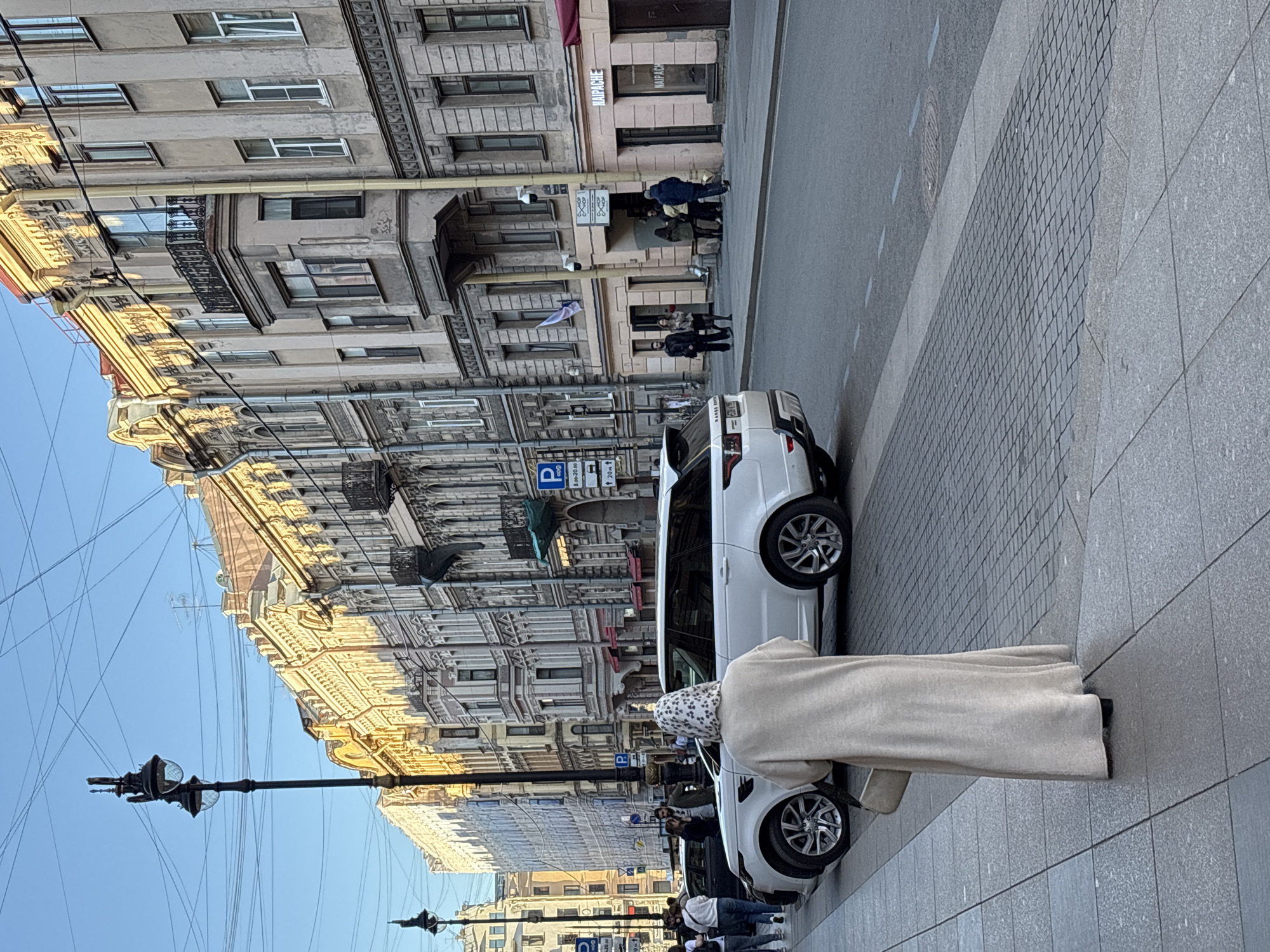 The scene is a street in Saint Petersburg, Russia. A white car is parked along the street, near a woman standing on the sidewalk. The woman wears a light-colored coat and a headscarf. There are several people walking on the sidewalk and near the building at the intersection. The buildings are ornate, with detailed facades and multiple windows. A black streetlamp stands on the sidewalk. There is signage visible on the front of the buildings.