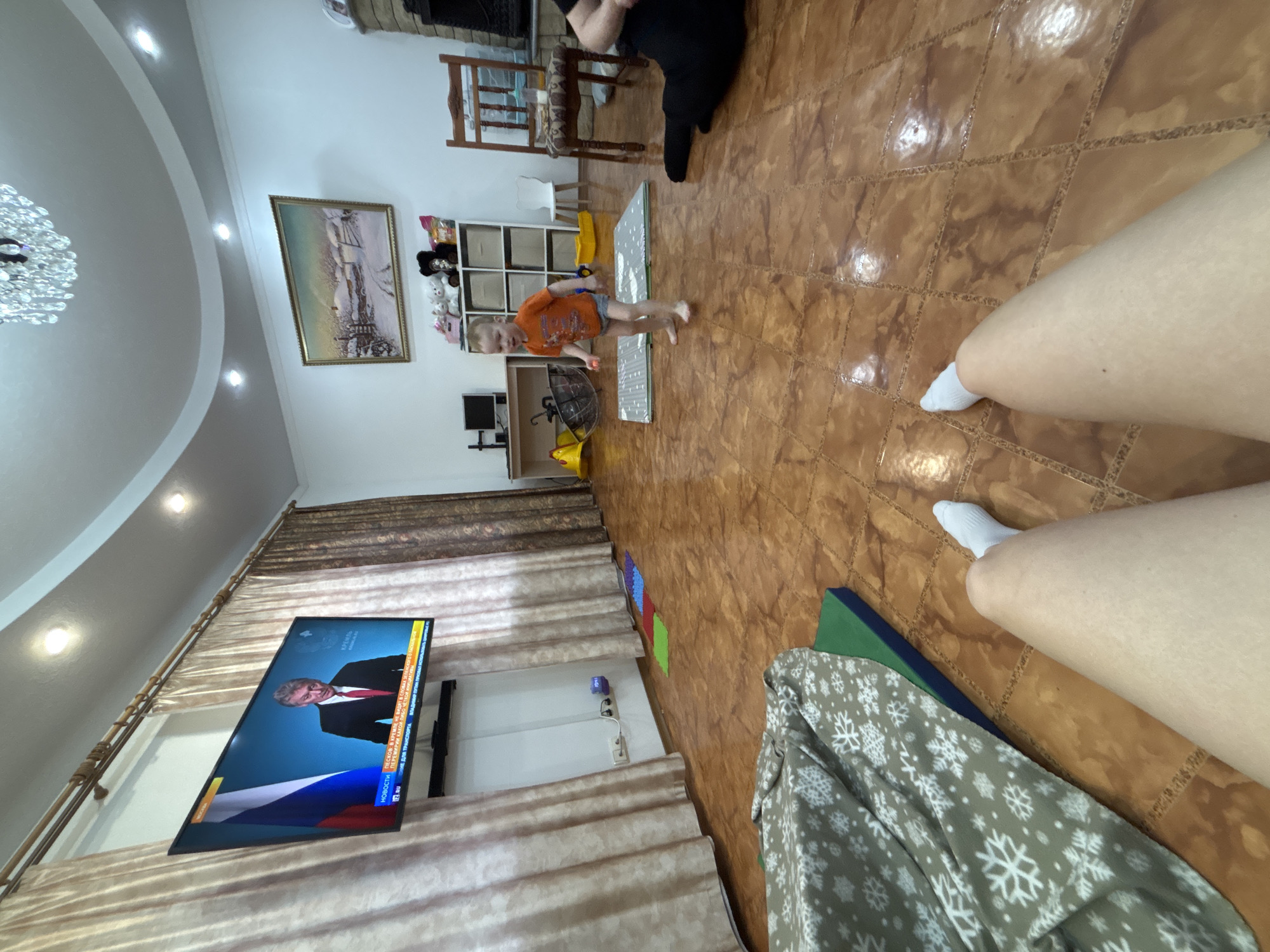 The image shows the interior of a residence located in Ferma, Russia. A young child stands on a grey foam mat in the middle of a tiled floor. A person's legs are visible in the foreground, possibly the photographer. There's a television on the wall displaying an image of Vladimir Putin, along with Russian text. Curtains cover the wall behind the television. To the left, a chandelier is visible on the ceiling. In the background, there's a fireplace, a painting on the wall, and a shelf with various items. A grey blanket with white snowflakes is on the floor.