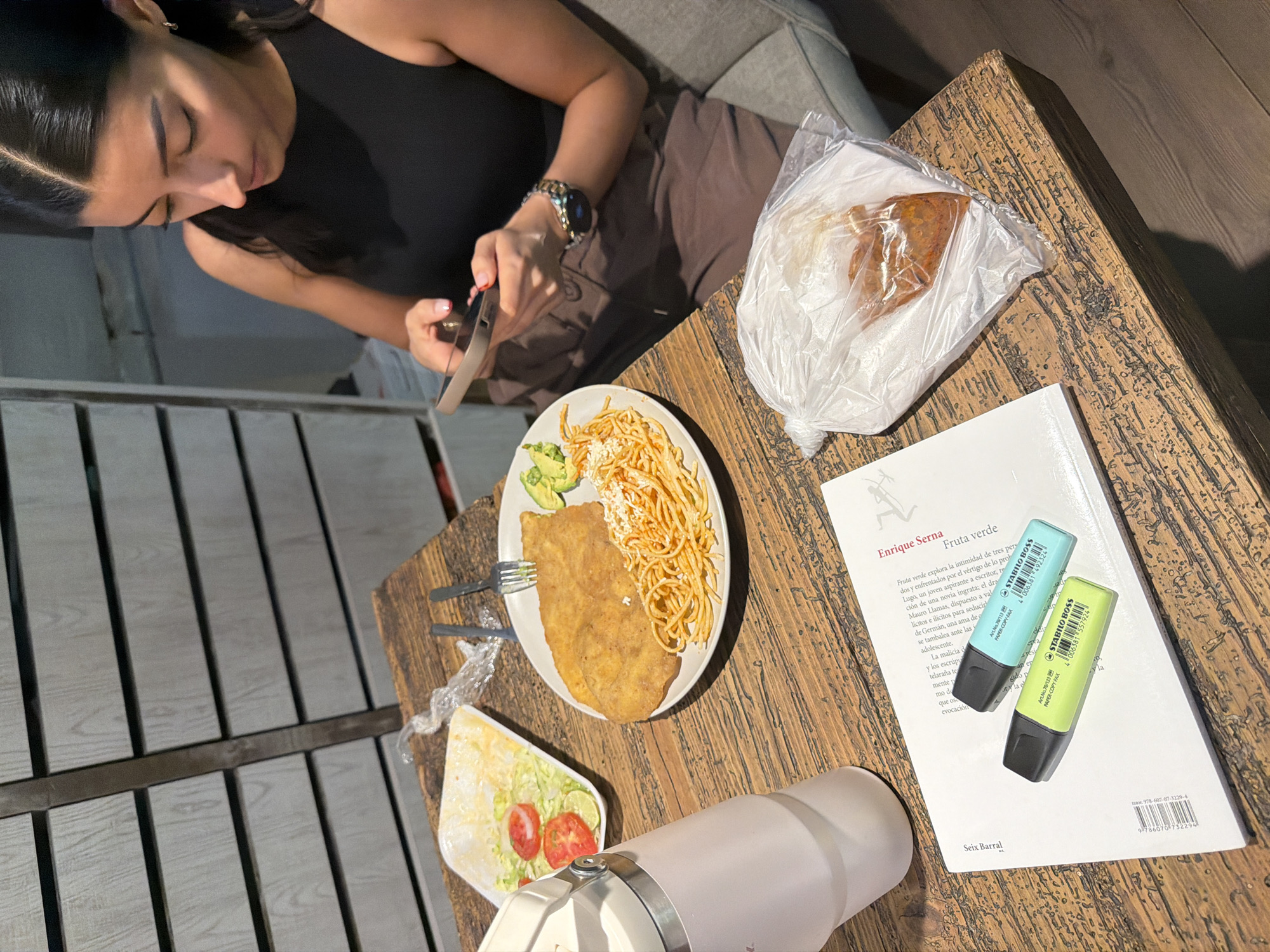 A woman sits on a sofa and looks at her phone while on a wooden table are arranged items of food, a book with two highlighters and a water bottle. The food consists of a plate with a piece of fried meat, spaghetti, and avocado, a container with salad, and a pastry in a transparent plastic bag. The book is titled "Fruta verde" by Enrique Serna. The highlighters are a light blue and light green Stabilo Boss. The water bottle is light pink and white. The location may be in Puebla, Mexico.