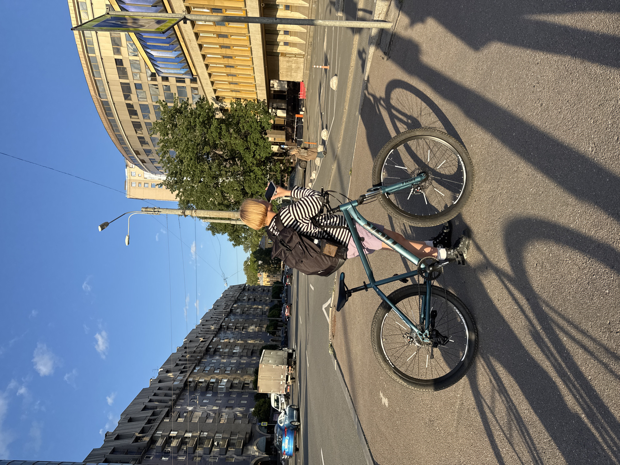 A person with blond hair, wearing a black and white striped long-sleeved shirt and light-colored shorts, stands next to a teal-colored bicycle on an asphalt sidewalk. The person is holding a mobile phone to their ear, appearing to be on a call. The bicycle has black tires, silver spokes, and a teal frame. In the background, a busy street with cars and trucks is visible. The street is flanked by tall, modern buildings. On the left side of the street is a long, gray, multi-story residential building with a distinctive angular facade. On the right side, a curved, light-colored building with many windows and a yellow-accented lower section stands. The sky is blue with a few white clouds, and the sun is casting long shadows of the person and bicycle across the sidewalk. The location is identifiable as Saint Petersburg, Russia, due to the distinctive architecture of the buildings, particularly the angular residential building and the curved administrative building, which are characteristic of Soviet-era modernist architecture found in the city. The presence of a pedestrian crossing sign further suggests an urban setting.