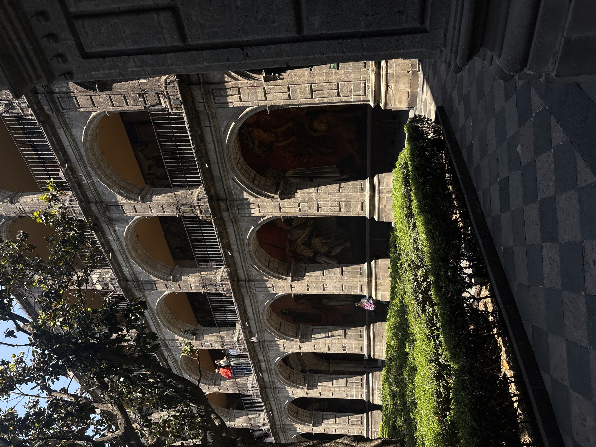 Here is a description of the image:
The image shows the Palacio Nacional (National Palace) in Mexico City, Mexico. The building has a facade with multiple arches, each containing frescoes. Some of the arches have wrought-iron bars. A tall green hedge runs along the base of the building. People are present on the balconies. A tree is visible on the left side of the frame, partially obscuring the building. The ground is paved with dark gray and black stones arranged in a checkered pattern. The image is shot from a slightly tilted angle, looking up at the building.