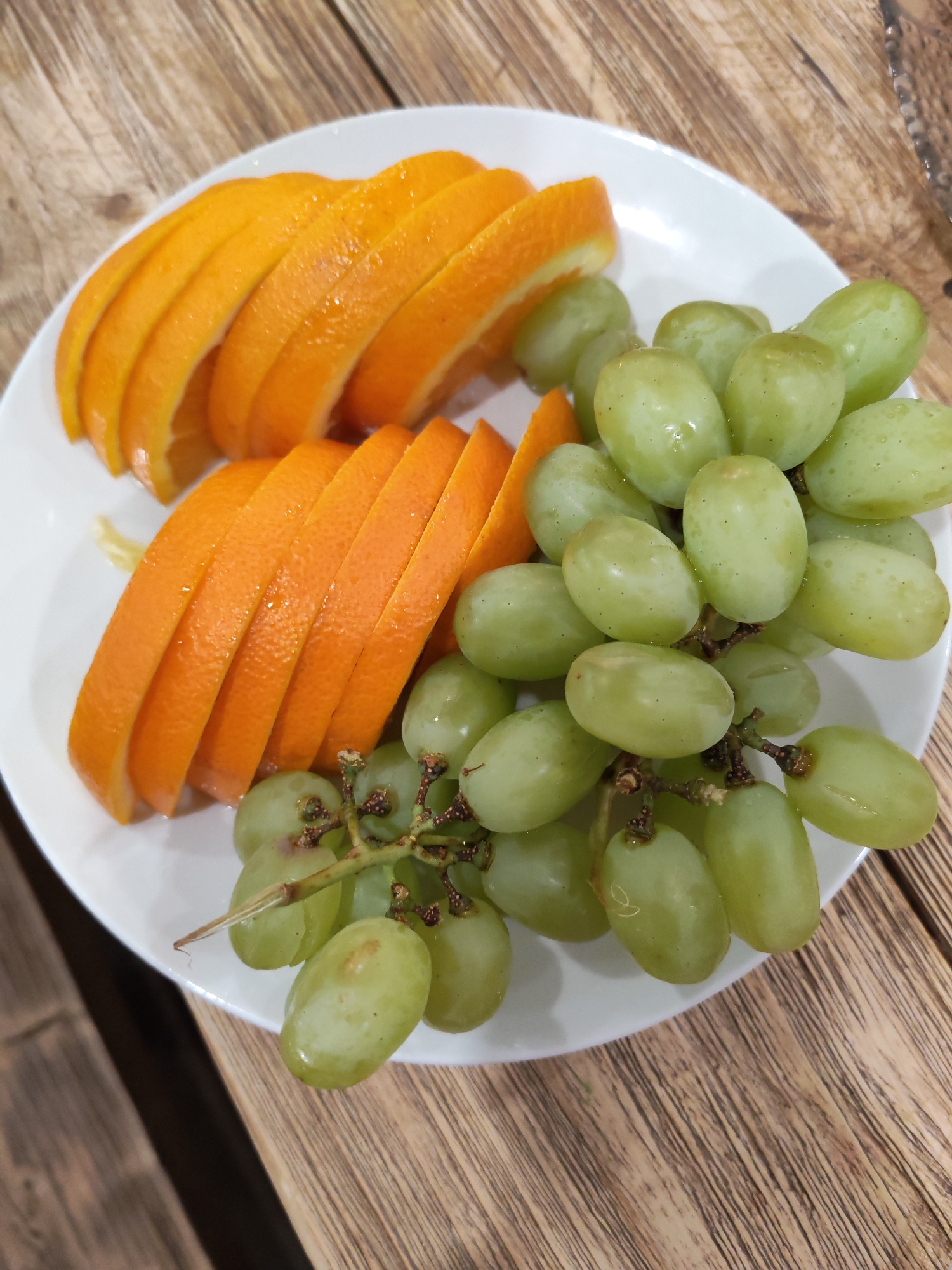 On a white plate, wedges of orange and bunches of green grapes are arranged. The plate sits on a wooden surface. The image was taken in Chaikovsky, Russia.