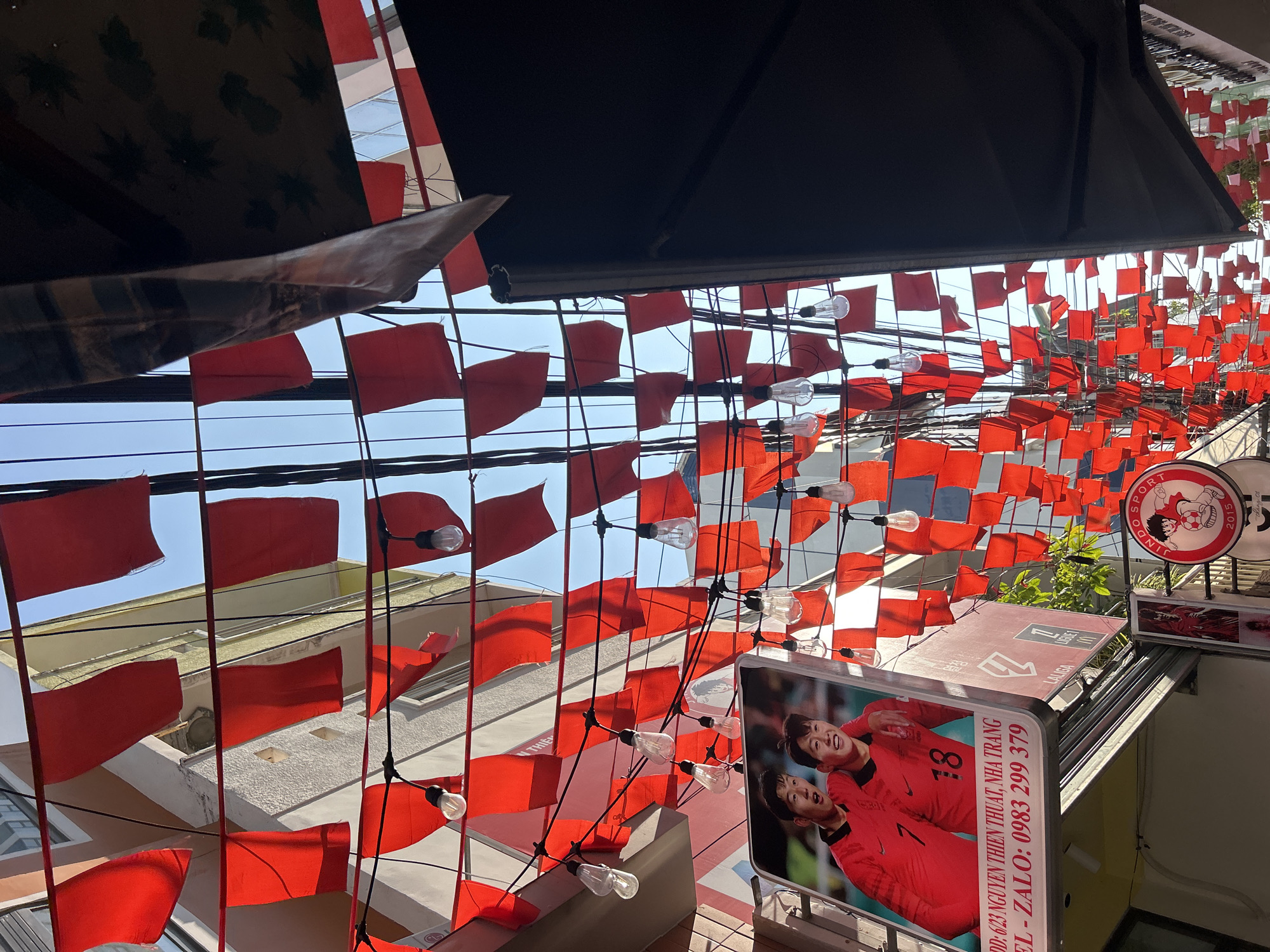 The image shows a street scene in Nha Trang, Vietnam, characterized by a dense display of small, red flags strung across overhead wires. The flags, likely decorations, are interspersed with strings of light bulbs. The view is angled upwards, capturing the sky and surrounding buildings.
Notable is the presence of signage. A sign bearing the numbers "7" and "18" and the text "DD: 6/23 NGUYEN THIEN THUAT, NHA TRANG" is visible. Other signs with sports-related logos and branding are also present.
The environment is urban, with buildings of varying heights flanking the street. The atmosphere appears to be daytime, with natural light illuminating the scene. The overall impression is a bustling commercial area with festive decorations.