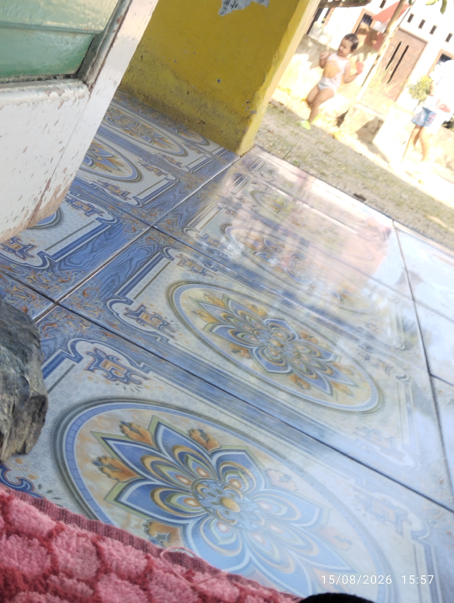 The image depicts an outdoor scene with tiled flooring in the foreground, featuring a blue and white floral pattern. To the left, a white door frame with peeling paint is visible, adjacent to a yellow wall. In the background, two children are present. One child, wearing a white t-shirt and diaper, stands in the sunlight. Another child, wearing a blue skirt, is partially visible on the right. The environment appears to be residential, with a house structure in the background. The timestamp "15/08/2026 15:57" is overlaid on the image. The location is identified as Tegal Rejo, Indonesia.