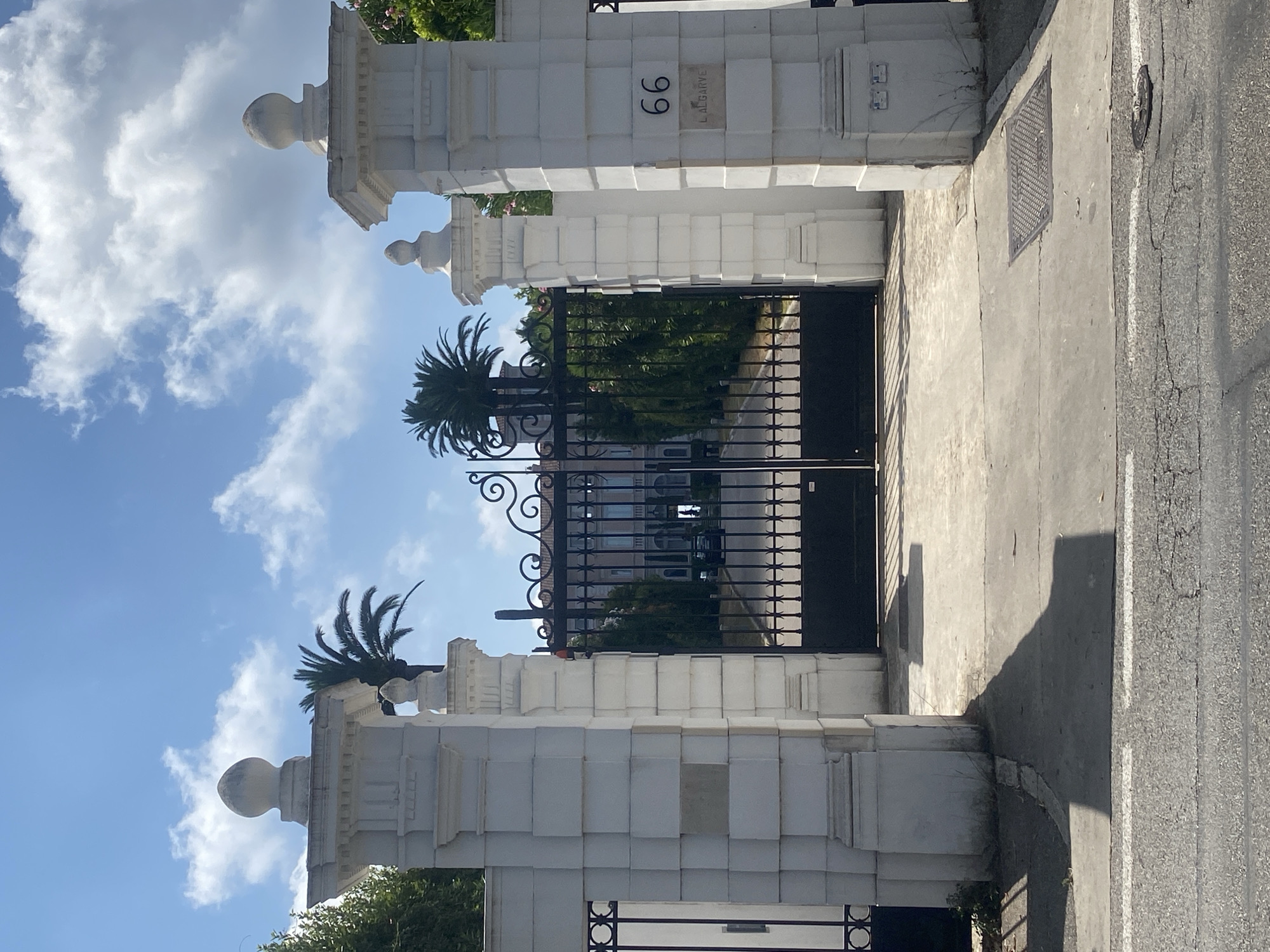 The image shows a grand entrance to a property on a sunny day in Antibes, France. The entrance is flanked by two large white pillars, one on the left and one on the right. The right pillar has the house number "66" and the name "L ALGARVE" inscribed on it. A black wrought-iron gate is centrally positioned, leading to a driveway lined with trees and leading to a building in the distance. Palm trees are visible behind the pillars, and the sky is blue with scattered white clouds. The foreground shows a paved road with white markings.