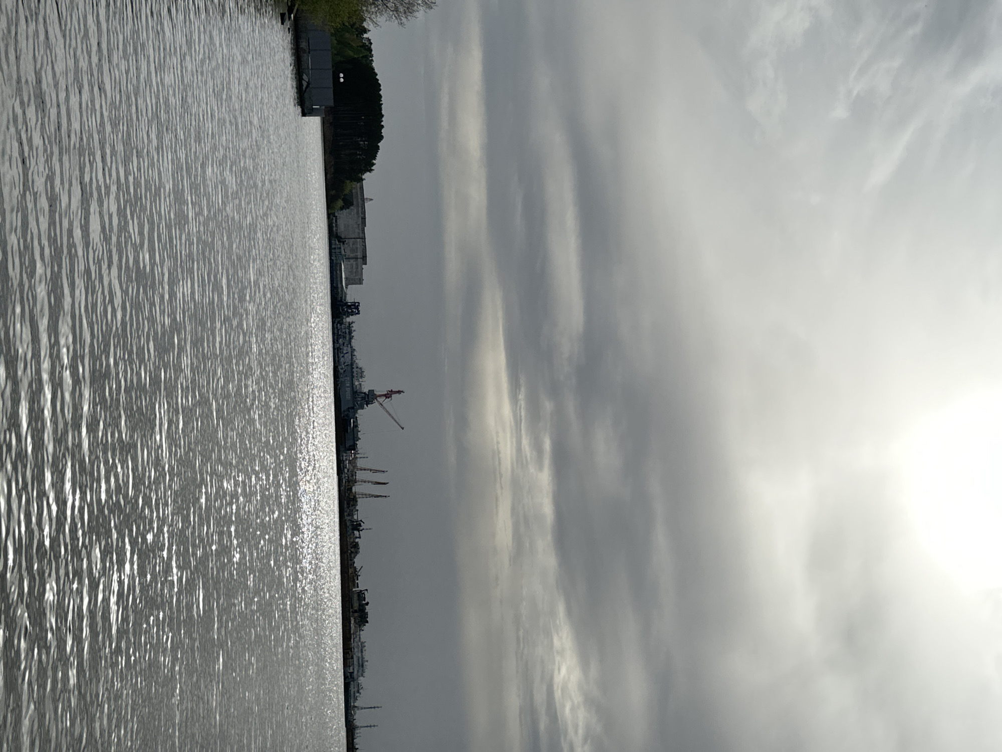 The image captures a body of water, possibly a river or bay, reflecting sunlight. Along the shoreline, a cityscape is visible with various structures, including industrial buildings, cranes, and ships, suggesting a port or industrial area. The sky is overcast with streaks of clouds, and the overall tone is gray and subdued. The location is the city of Unknown, Unknown.