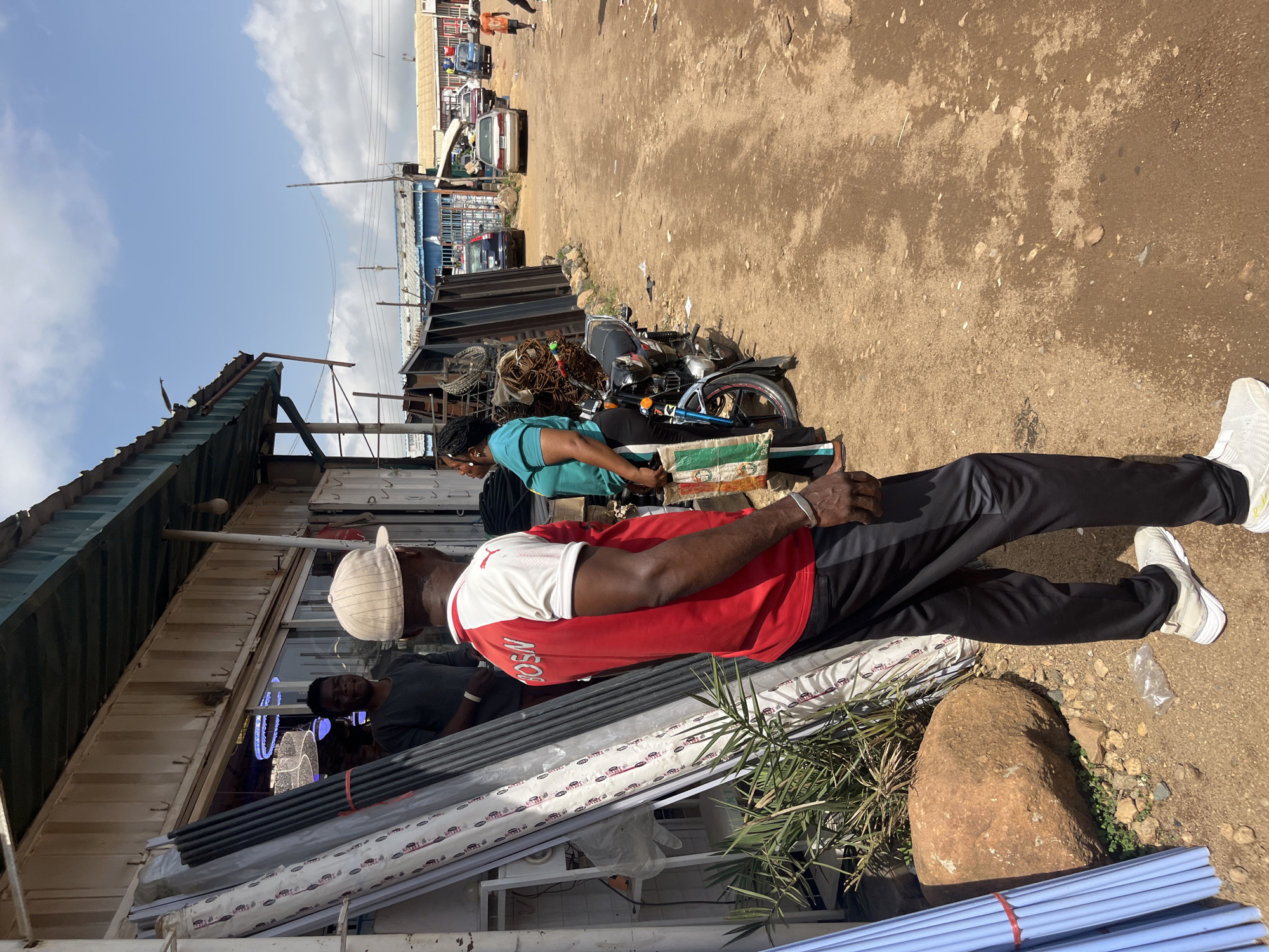 The image depicts an outdoor scene in Gura Top, Nigeria, likely during the day under a partly cloudy sky. The foreground is dominated by a man wearing a red and white t-shirt, black pants, and a white cap. He is facing away from the camera, and his left hand is resting on a stack of long, light-colored pipes bundled with red ties. To his right, a large rock is visible. Behind him, another man in a dark shirt is partially visible, also looking away.
In the midground, a woman in a turquoise shirt is seated on what appears to be a bag, leaning against a motorcycle. The motorcycle is parked on a dirt ground, which also has scattered debris. Behind her, the scene opens up to a marketplace or industrial area with various structures, including corrugated metal roofs and stalls. Several vehicles, including cars and a van, are parked in the background, suggesting a busy commercial area. Power lines are visible stretching across the scene, and some buildings have metal sheeting.
To the left of the image, a building with a corrugated metal roof and a white structure attached to it is prominent. The overall impression is that of a bustling outdoor market or informal business district. The lighting suggests it is daytime, and the weather appears to be clear or partly cloudy. No specific emotions are discernible from the subjects, as most are not facing the camera. Text is not clearly legible on any visible objects.