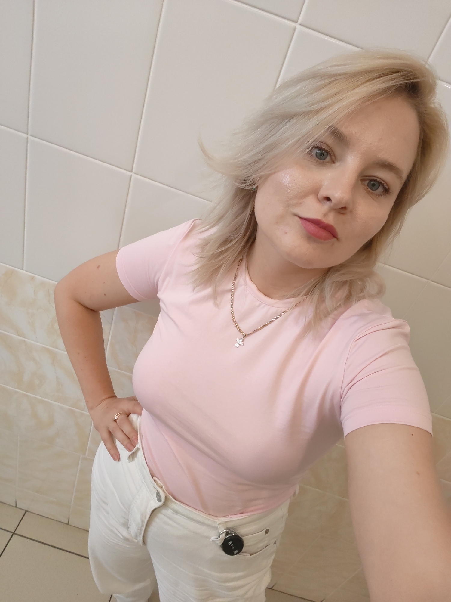 A woman with blonde hair and blue eyes stands in front of a tiled wall. She is wearing a pink t-shirt and white pants. She is looking at the camera and has her hand on her hip. The background appears to be a bathroom. The image was taken in Izhevsk, Russia.
