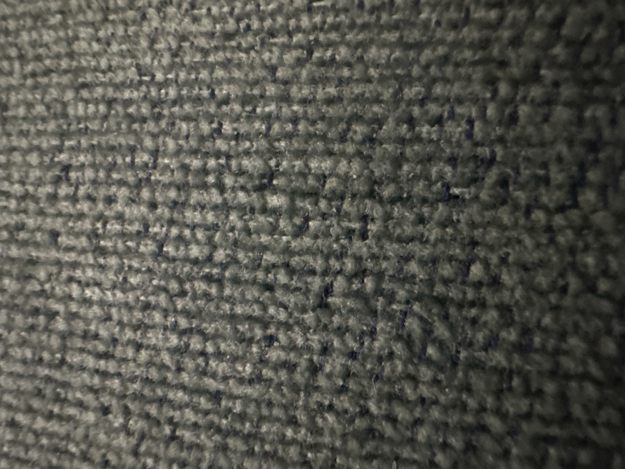 The image depicts a close-up view of a grey, looped carpet. The lighting is uneven, with some areas appearing darker than others. The individual loops of the carpet fibers are visible and uniformly spaced. There is no discernible location information or other subjects or objects present.