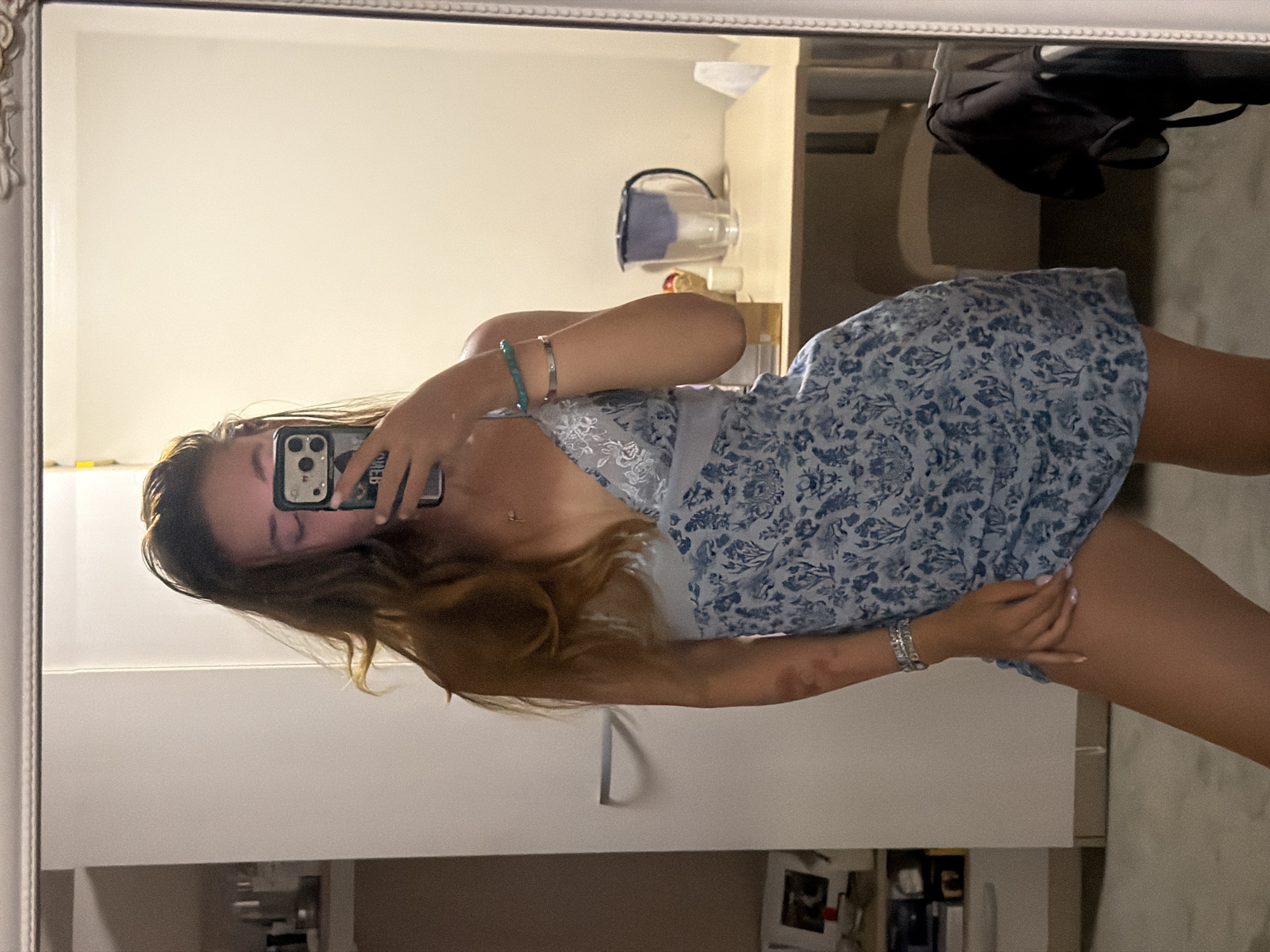 A young woman with long, light brown hair is reflected in a large, ornate mirror, taking a selfie with her phone. She is wearing a light blue slip dress with a floral pattern. The dress has a lace trim on the V-neckline and thin straps. She is also wearing two bracelets on her left wrist, one silver and one teal. Her right arm is bent at the elbow and her hand is resting on her thigh. She has a small necklace with a pendant around her neck.
The background of the reflection shows a room. To the left, there is a tall shelf unit with various items. In the center, there is a light-colored desk with a water filter pitcher and a small bowl with fruit on it. Behind the desk, a chair is partially visible. To the right, a black garment is hanging on the mirror frame. The floor is a neutral gray. The overall lighting suggests an indoor environment, likely a bedroom or dressing area.
The image does not contain any information that allows for the identification of the city as Sofia, Bulgaria.
