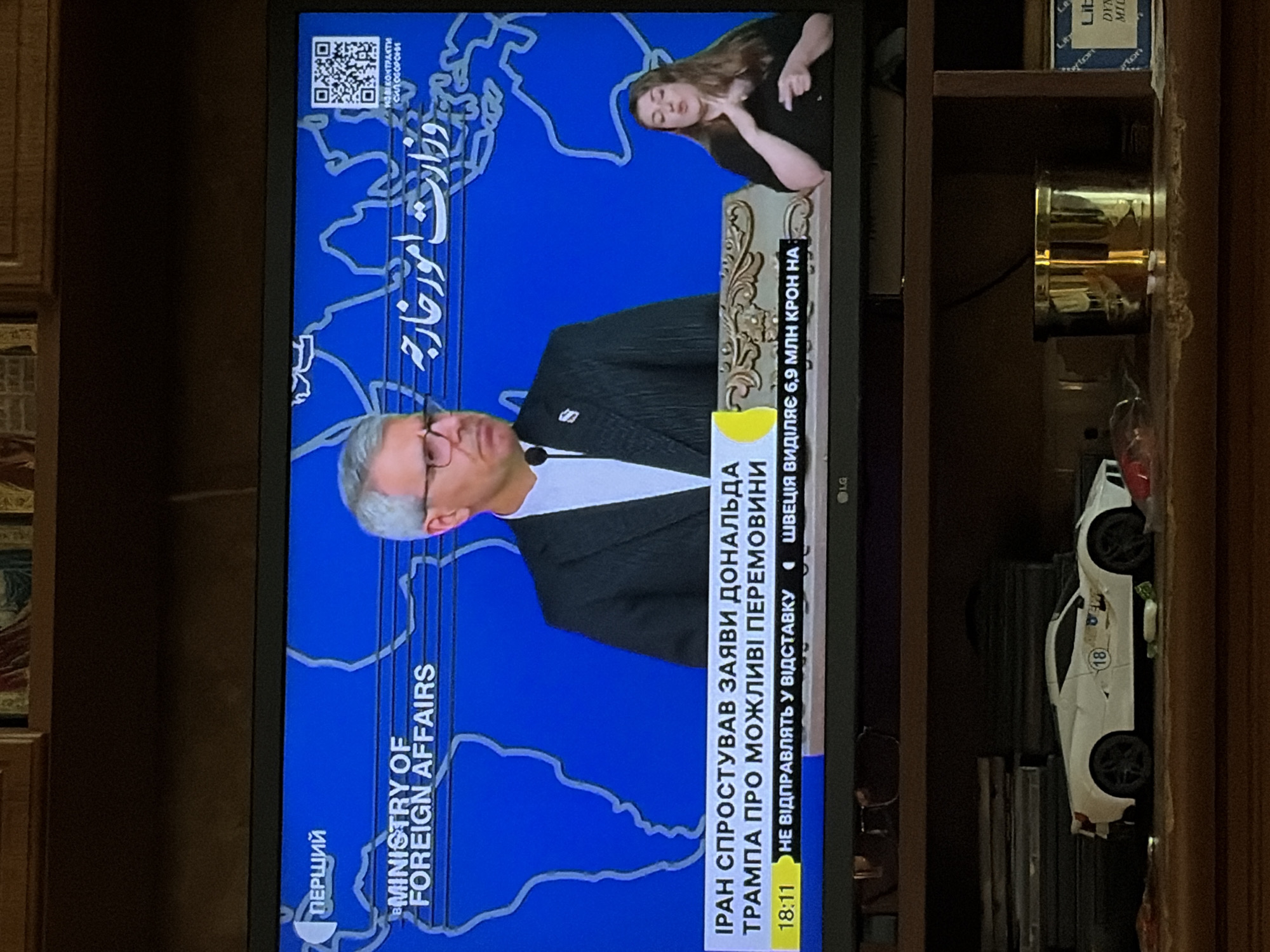 A television screen displays a news broadcast with a male news anchor, a sign language interpreter, and text overlays. The background of the broadcast features a world map graphic. Text in Ukrainian and Arabic is visible on the screen. The Ukrainian text translates to "IRAN DENIES DONALD TRUMP'S STATEMENTS ABOUT POSSIBLE NEGOTIATIONS". Below this, "18:11" is displayed alongside other Ukrainian text. The Arabic text reads "وزارت امور خارجه" which translates to "Ministry of Foreign Affairs". The screen is positioned on a shelf in front of a bookshelf. Below the television, on the same shelf, are several objects including a white toy sports car with the number "18" on its side, a gold-colored cylindrical container, and part of a blue box with white text. The overall scene appears to be an indoor environment.