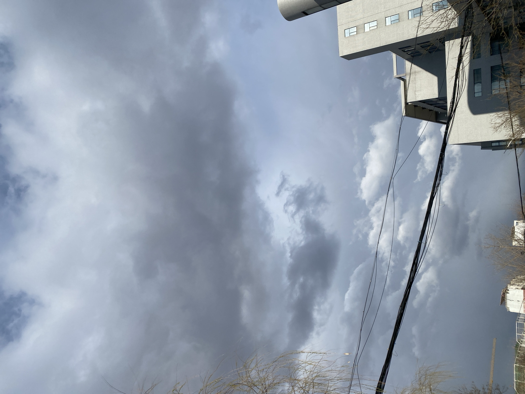 The image captures an upward view, likely in Zhongtun, China. The sky is filled with a mix of dark, heavy storm clouds and patches of lighter, brighter clouds, indicating unsettled weather. A modern, grey-toned building dominates the upper right corner, featuring rectangular windows. Wires, presumably for power or communication, stretch across the scene, intersecting the building and the sky. Bare branches of trees are visible in the lower portion of the image, hinting at the time of year, likely late autumn or winter. No people are visible.