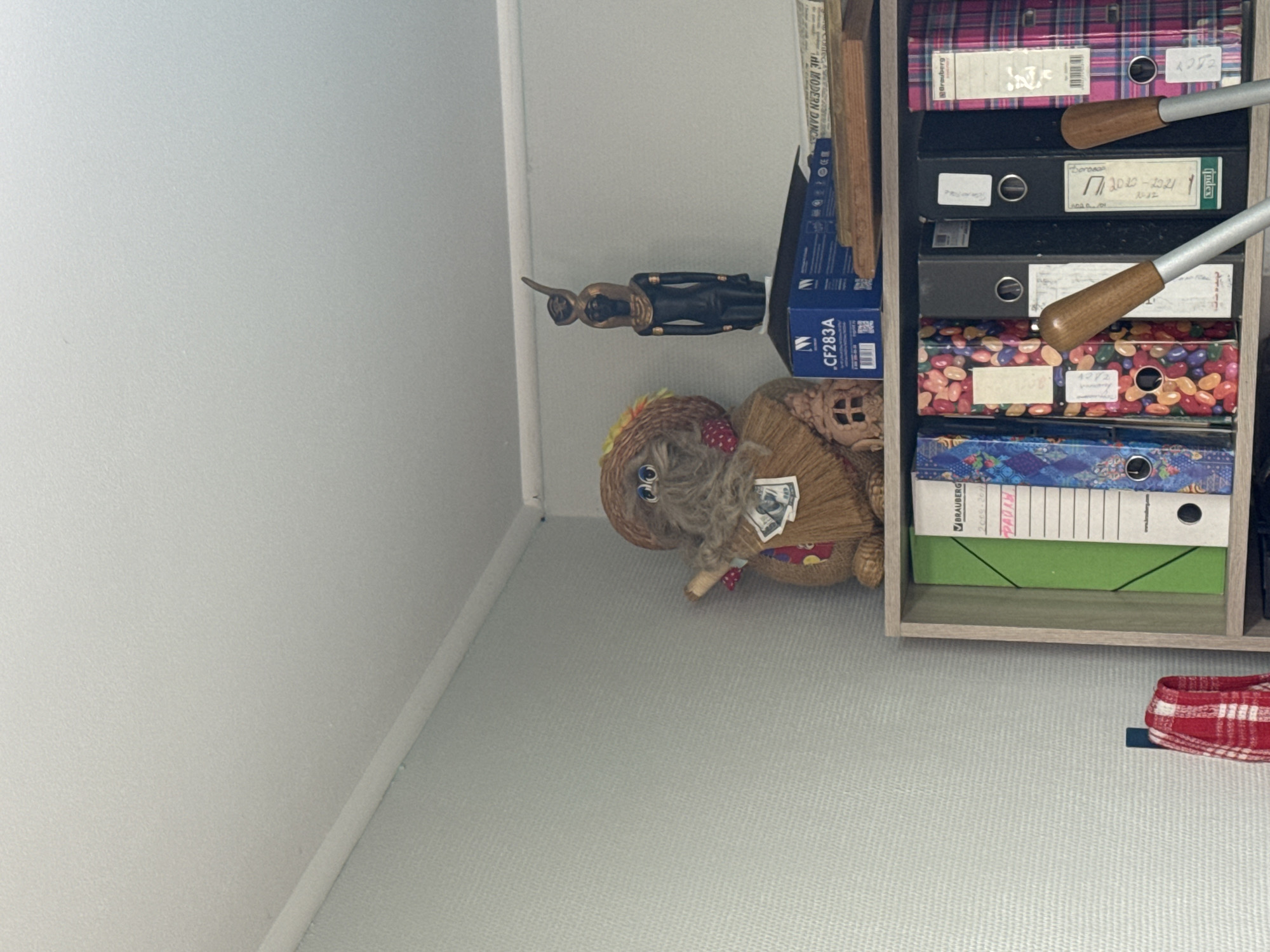 The image displays the interior of a room, likely a shelf or cabinet, in Chaikovsky, Russia. On the top shelf, a straw doll figure with large blue eyes and a straw hat is positioned to the left. To its right stands a dark-colored figurine of an Egyptian deity, possibly Isis, on top of a blue box with the label ".CF283A". Additional items, including what appears to be paper or books, are visible behind the figurine.
The lower shelf holds several binders. From left to right, there is a binder with a blue and patterned cover, followed by one decorated with jellybeans. Next are two black binders, and to the far right is a binder with a pink and black plaid pattern. Two wooden handles of cleaning tools, likely brooms or mops, are partially visible in the foreground, obscuring some of the binders. A red and white checkered cloth is hanging on a hook to the left of the lower shelf. The overall environment suggests an organized storage space within a residential or office setting.