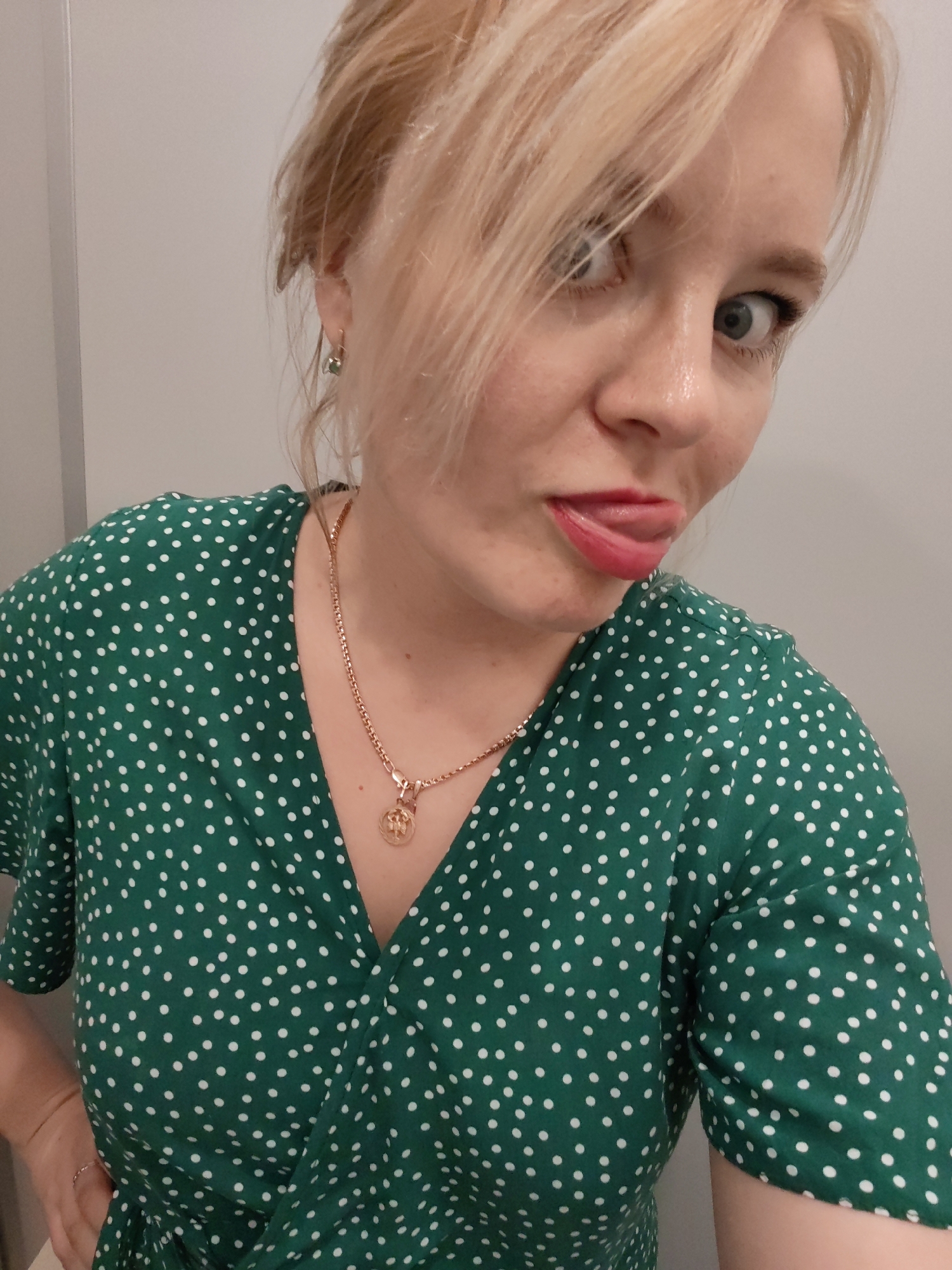 A woman with blond hair, pulled back loosely, is posing for a close-up selfie. She is sticking her tongue out and making a slightly cross-eyed expression. She is wearing a green dress with white polka dots and a gold necklace with a pendant. The background is a plain white wall. There is no evidence to suggest that the image was taken in Izhevsk, Russia.