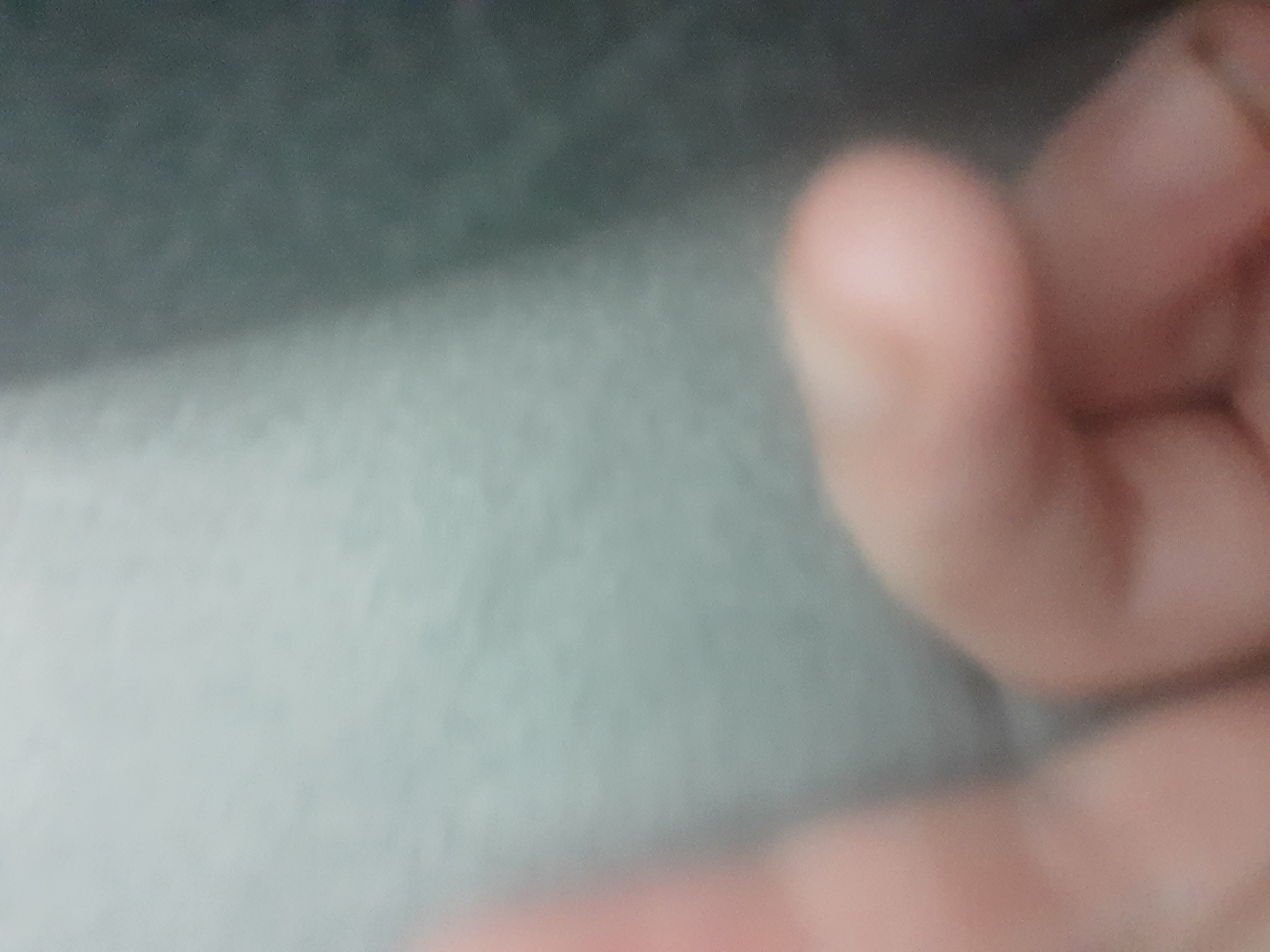 The image is a blurry, close-up shot of a person's hand. The background is out of focus and appears to be a textured surface in shades of gray and light blue. There are no identifiable landmarks or context that would allow for location determination within Liubakhy, Ukraine, or any other specific location. The image is too underexposed and lacking in detail to provide a forensic description of a scene.