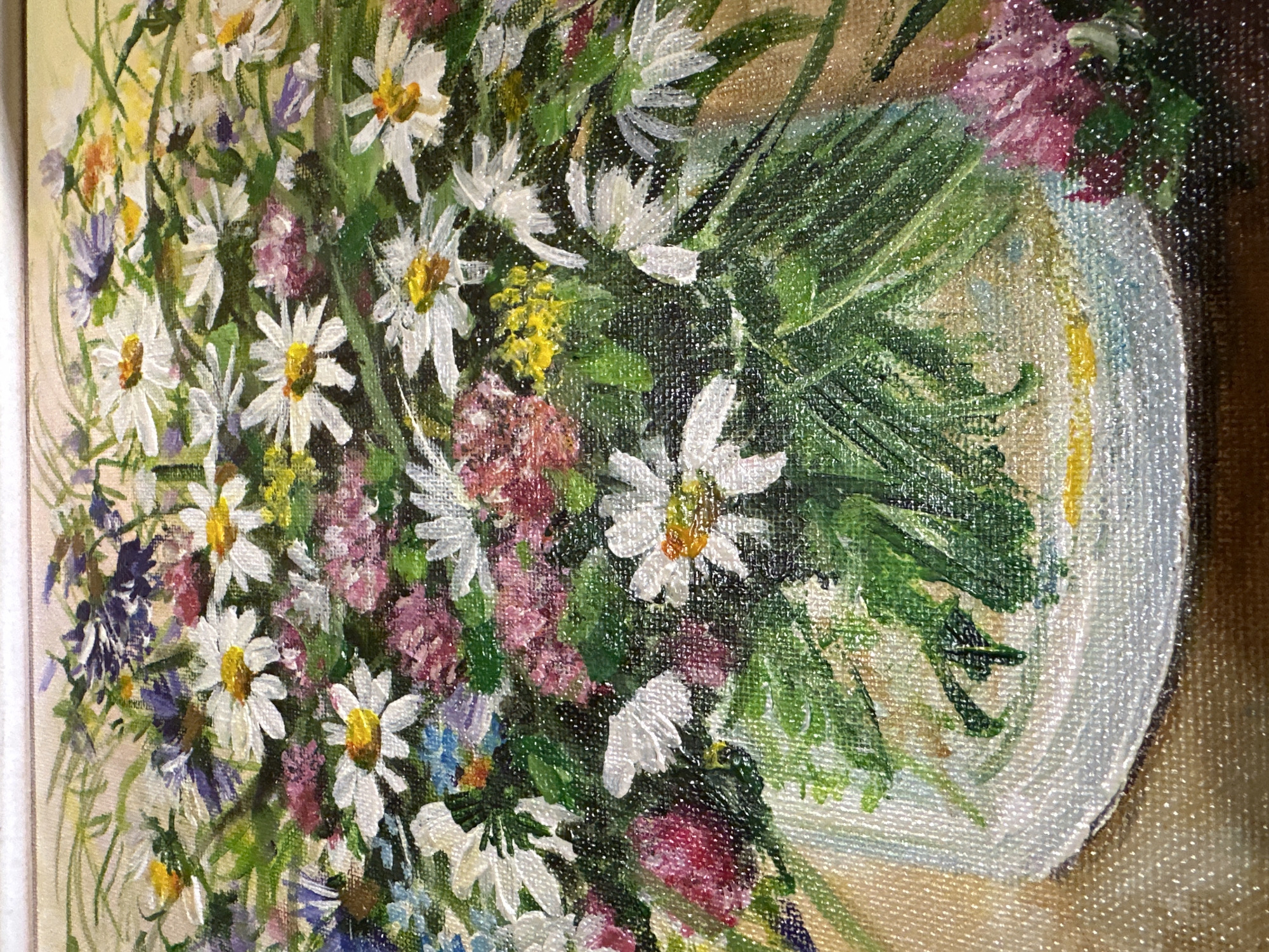 The image is a close-up, overhead shot of a painting depicting a vibrant bouquet of wildflowers in a white vase. The flowers are a mix of white daisies with yellow centers, small pink and purple flowers, and delicate blue blossoms, all surrounded by lush green foliage. The painting's texture, likely oil on canvas, is visible, with visible brushstrokes. The vase is simple and white, with a textured rim, and sits on a surface that appears to be a light brown tablecloth or tabletop. The background is a soft, muted beige. The painting is lit from above, casting subtle shadows. There are no people or animals present in the painting. The image does not provide any information that would allow for its location in Sergeevka, Russia.