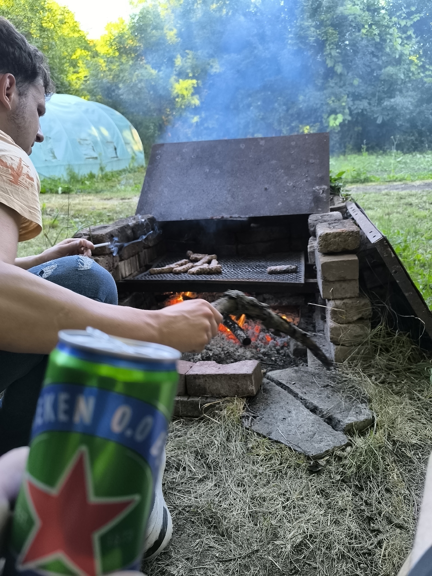 An outdoor scene shows a person in the foreground holding a green Heineken 0.0 can. In the mid-ground, a person is sitting next to a brick barbecue grill, smoking a cigarette and flipping sausages on the grill with a stick. The grill is made of bricks and has a metal grate. Coals are glowing beneath the sausages. Smoke rises from the grill and mixes with the blue smoke from the cigarette. In the background, there is a greenhouse and trees. The ground is covered with grass and dry vegetation. The scene appears to be in a backyard or park setting, possibly in Sofia, Bulgaria, given the context of the question.