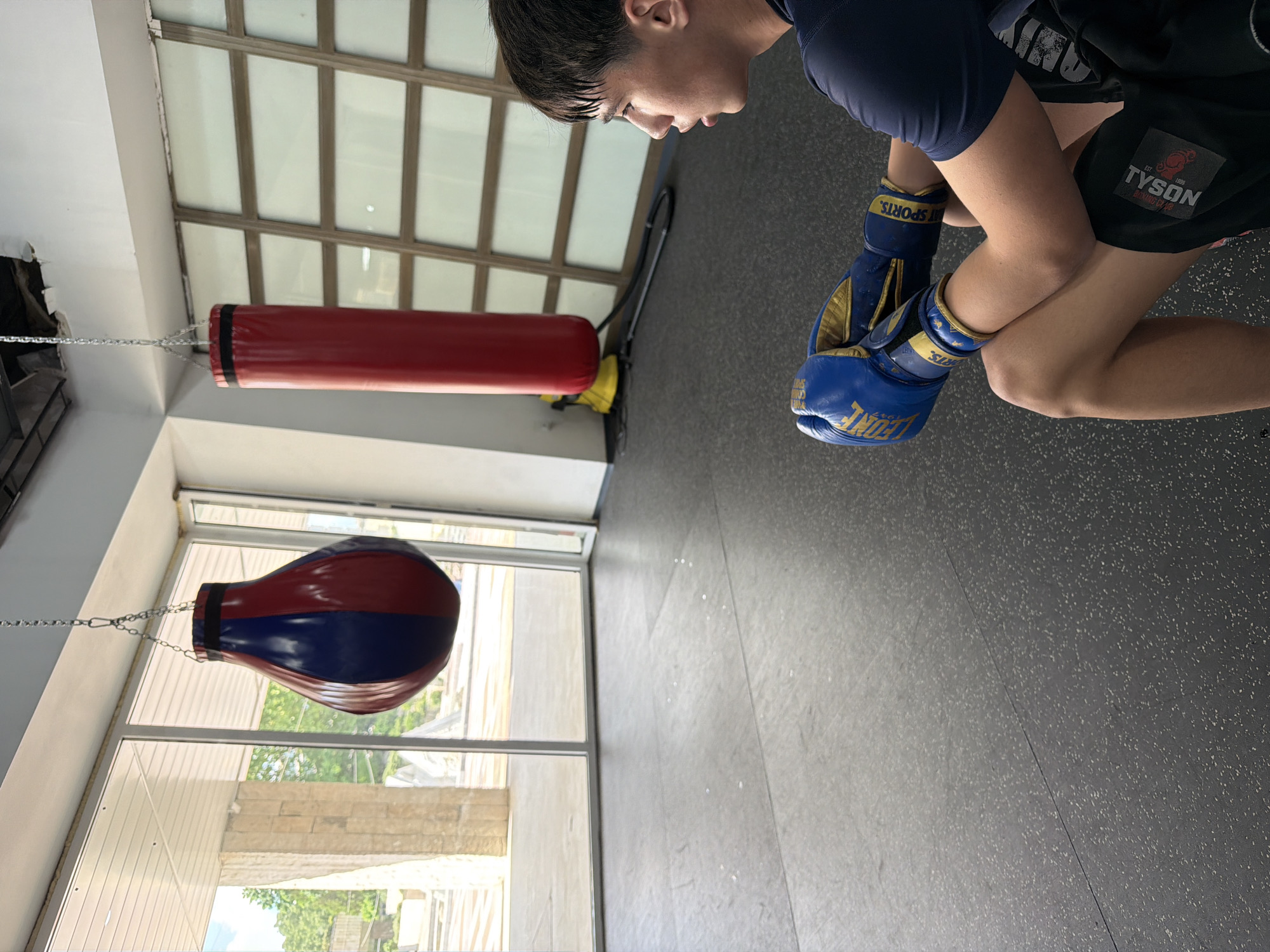 A boxing gym interior featuring a person with boxing gloves and boxing bags. A youth with dark hair and dark clothing, wearing blue and gold boxing gloves, is visible in the upper right corner of the frame. The lower part of the frame showcases a red and blue speed bag hanging in front of a window, and a long red punching bag is positioned in the background. The gym has a grey floor and a window showing a cityscape, potentially placing the location in an urban setting, potentially Blagoevgrad, Bulgaria.
