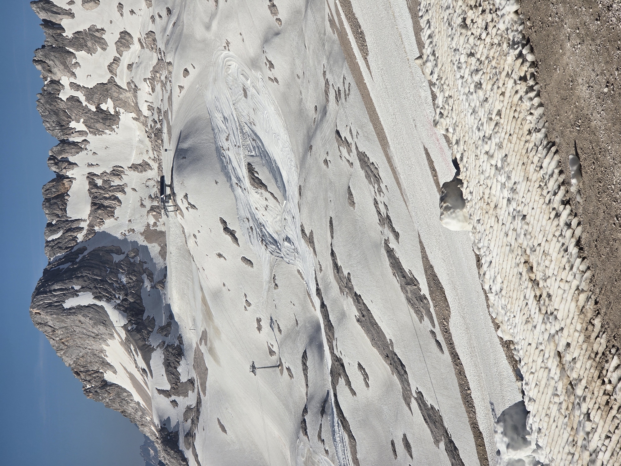 This is a high-angle, long-shot, eye-level outdoor photograph of a snow-covered mountain landscape under a clear blue sky. The image depicts a vast, rocky mountain range with significant snow cover, indicating a high altitude. The terrain is characterized by steep slopes, patches of exposed rock, and areas of compacted snow with visible tracks, suggesting it might be a ski resort or a hiking area.
In the mid-ground, a ski lift station with a dark-colored roof and supporting structure is visible, situated on a snowy ridge. A ski lift cable extends from this station. Further down the slope, another ski lift pylon stands in the snow. The snow surface is not uniform; there are areas of smooth snow, as well as textured patches, possibly from previous skiing activity or natural snow formations.
In the foreground, a section of the terrain is covered with a textured, granular material, likely gravel or dirt, interspersed with rows of white, somewhat rectangular blocks. These blocks appear to be made of packed snow or ice, arranged in a pattern that creates a ridged surface. A large, irregularly shaped mound of snow rests on the left side of this foreground feature.
The background is dominated by the jagged peaks of the mountains, with snow clinging to their upper reaches and filling crevices. The overall impression is one of a remote, mountainous environment, likely a tourist destination known for winter sports or alpine scenery. The location is a high-altitude area near Garmisch-Partenkirchen, Germany, possibly the Zugspitze region, known for its glaciers and ski resorts. No people are visible in the image.