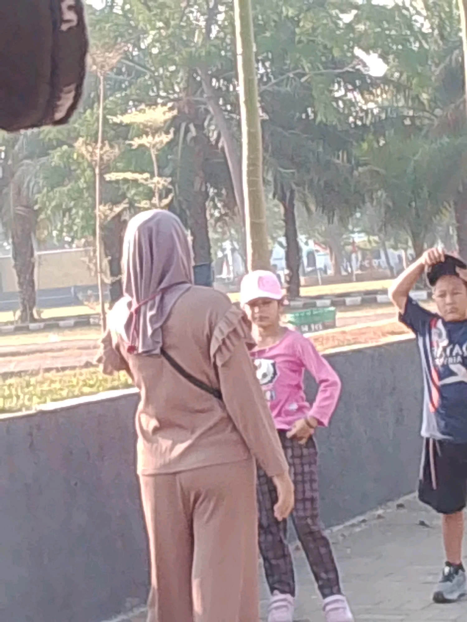 In Probolinggo, Indonesia, three individuals are observed outdoors. In the foreground, a person wearing a beige hijab and matching outfit stands facing away from the camera, with a black bag slung across their body. To their right, a young girl in a pink long-sleeved shirt and plaid pants, with a pink cap, looks towards the left. Further to the right, a young boy in a dark t-shirt and black shorts, wearing a cap on his head, is seen adjusting his cap with his right hand. The setting appears to be a park or outdoor recreational area, with trees and greenery visible in the background, along with paved pathways and a low wall. The lighting suggests it is daytime.