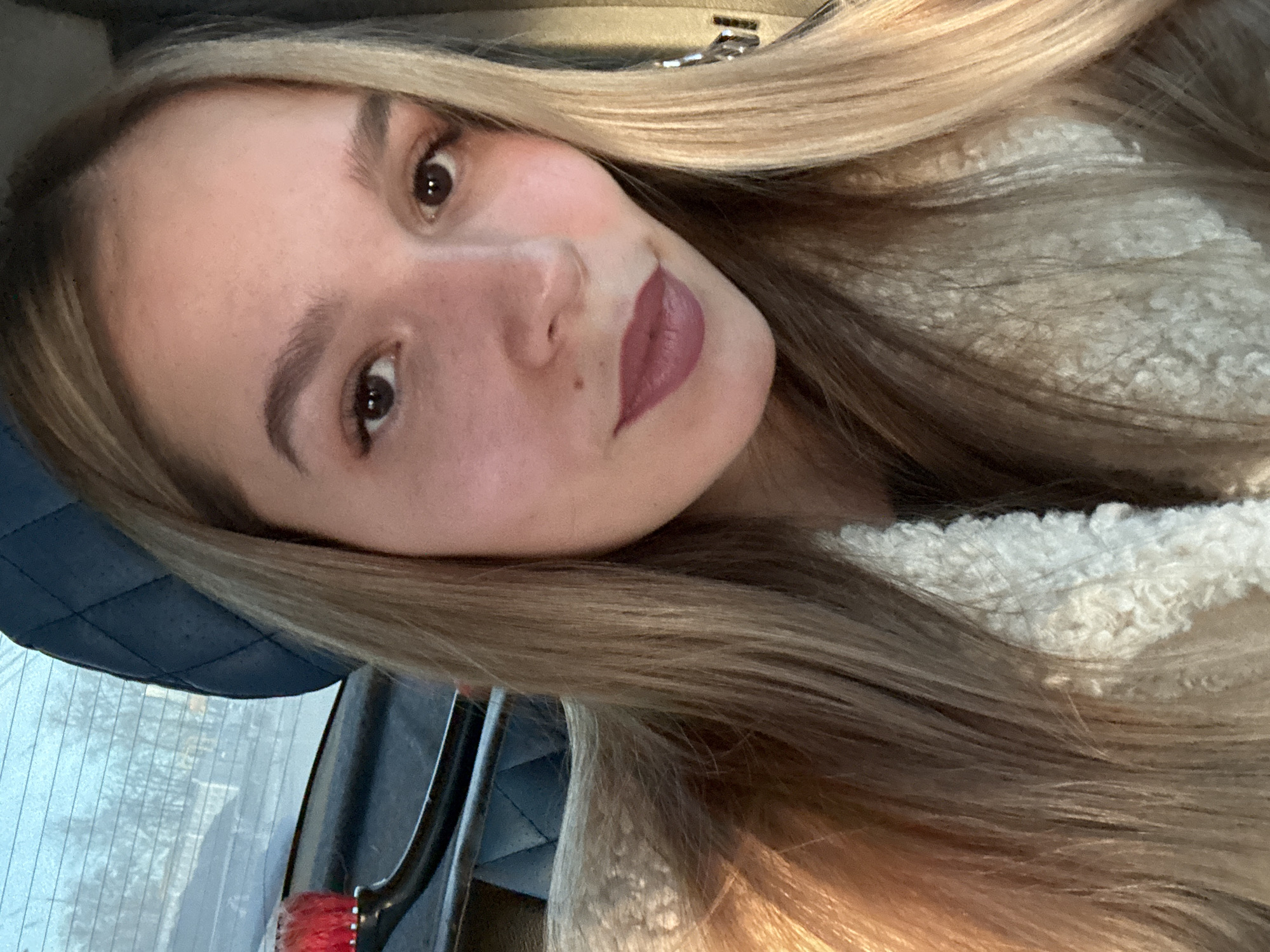 A young adult female is depicted in a close-up, seated inside a vehicle in Chaikovsky, Russia. The subject has fair skin, long straight blonde hair, and dark brown eyes, looking directly at the camera. She wears matte, dusty rose-colored lipstick, and a small mole is visible on her right cheek, inferior to her right eye. She is dressed in a cream-colored, textured, possibly sherpa-style garment. The background reveals the interior rear of the vehicle, with a dark blue quilted headrest partially visible to the upper left of the subject's head. A dark metallic bar extends horizontally across the mid-left portion of the frame, to which a small, red, brush-like or pompom-like object is attached. Through the vehicle's rear window, a blurred outdoor environment consisting of a light blue sky and faint tree line is visible.
