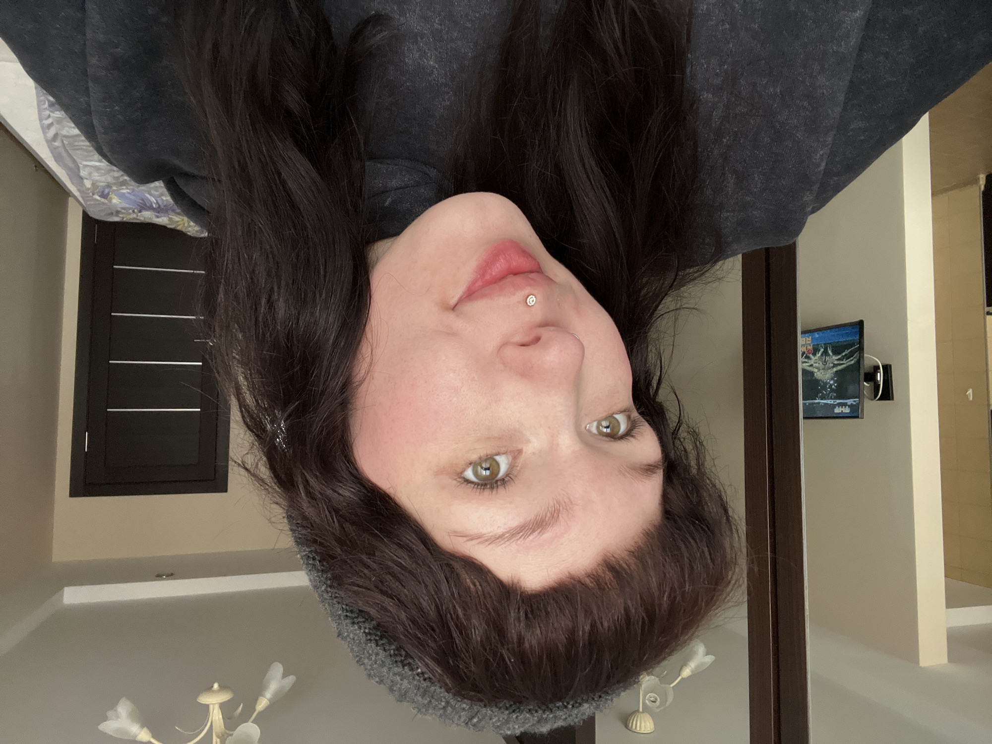 A selfie of a woman with green eyes and dark hair is oriented upside down. She has a piercing above her lip on the right side of her face. She is wearing a grey hoodie and a grey beanie. In the background, there's a TV screen on a wall, a dark-colored door or window, a chandelier, and a hallway. No information related to the location in the city of Kazan, Russia, is discernible in the provided image.