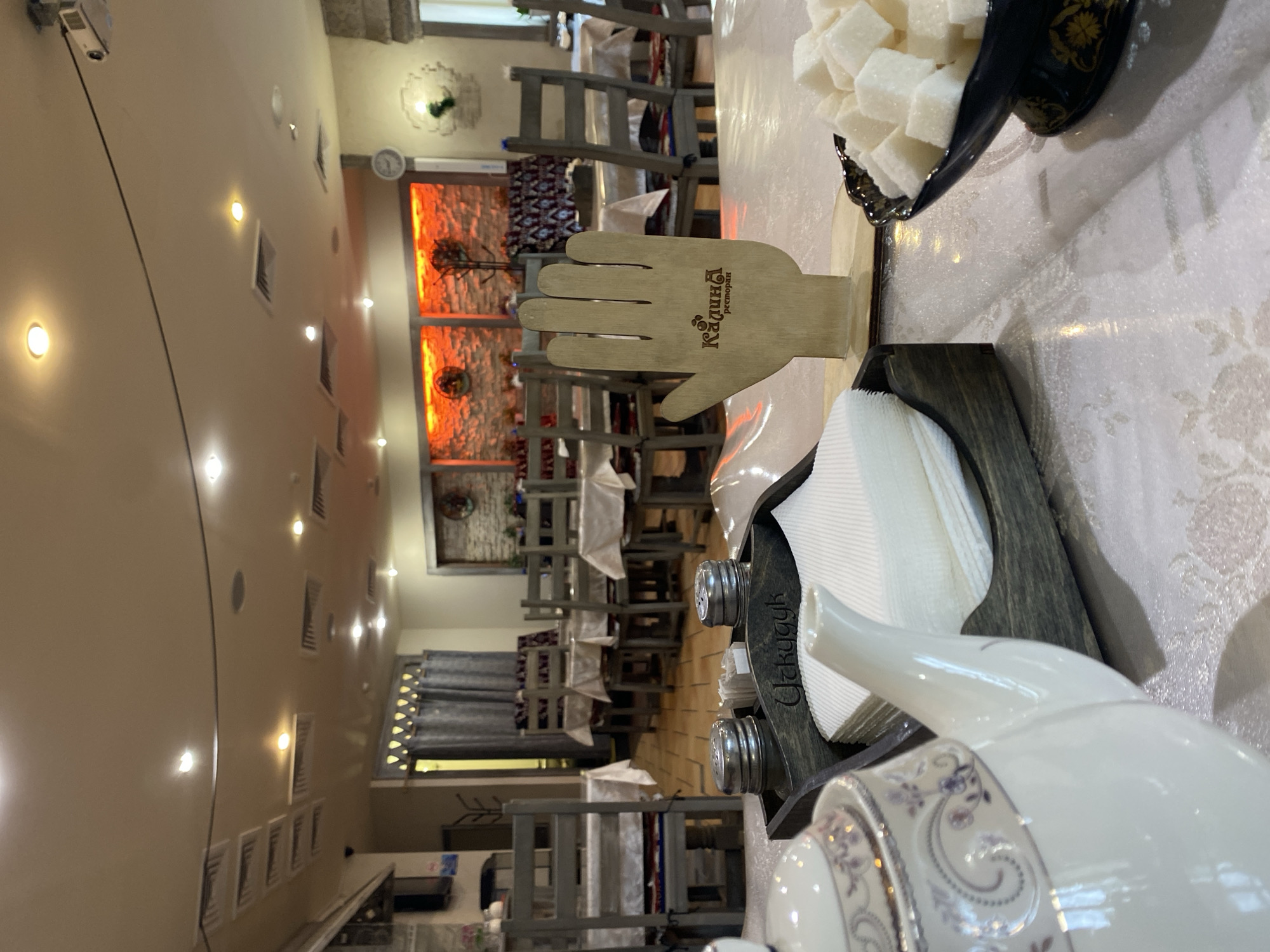 The image displays the interior of a restaurant or cafe, viewed from a table in the foreground towards the main dining area. The establishment appears to be empty of patrons, suggesting it is either closed or prepared for service.
On the table in the immediate foreground, a white ceramic teapot adorned with subtle floral patterns is visible. Beside it rests a dark wooden caddy containing a stack of white paper napkins and two metallic shakers, likely for salt and pepper, with the word "Ozkuygy" faintly inscribed on it. A black bowl filled with white sugar cubes is also present. A distinctive wooden stand, shaped like a human hand, prominently features the text "Калина ресторан" (Kalina restaurant), identifying the venue.
The dining area beyond is furnished with numerous wooden tables and chairs, many of which are covered with white tablecloths. The walls are a light, neutral color, illuminated by multiple recessed circular lights and fitted with rectangular air vents. In the mid-ground, decorative wall panels emit a warm, orange-red glow, contributing to the ambiance. Further in the background, a projector is mounted on the ceiling, windows are covered by dark curtains, and a patterned rug or tapestry hangs on a wall. The scene depicts a well-arranged and inviting dining space located in Ryazan, Russia.