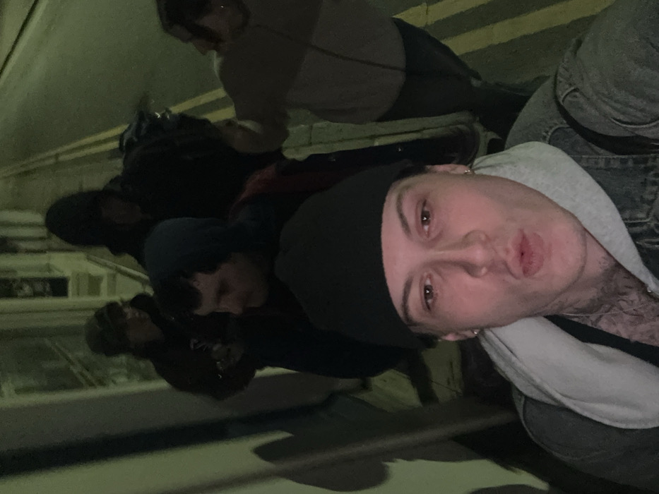 This image captures a nighttime street scene in Deal, United Kingdom, featuring five individuals. In the immediate foreground, a male with light skin, wearing a black beanie, a grey hooded sweatshirt, and a denim jacket, is presenting a pursed-lip expression directly to the camera, consistent with taking a selfie. He has visible earrings and tattoos on his neck/upper chest. Behind him and to his left, another male in a dark hooded top is partially visible. Further back, three more individuals are present: one on the left wearing a dark baseball cap, a central figure in a dark hooded top carrying a backpack, and a fifth individual on the right wearing a lighter two-toned top and darker trousers. The scene is illuminated by an artificial light source, casting shadows on a wall to the left. A paved surface with double yellow lines, indicative of a public road in the UK, is visible on the right.