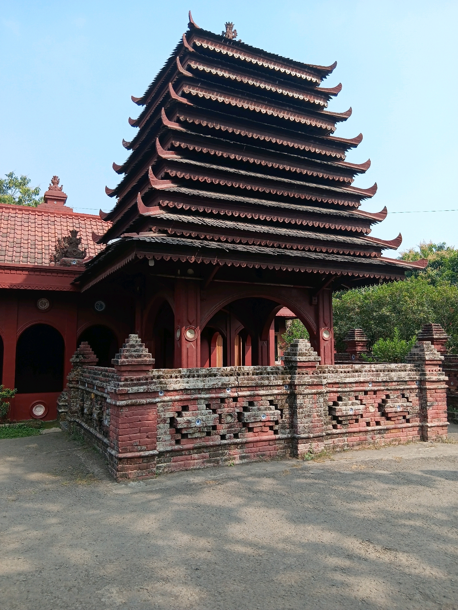 This is a full-shot, eye-level, outdoor photograph of the Menara building at Sunyaragi Archaeological Park in Cirebon, Indonesia. The building is a multi-tiered, pagoda-like structure with a dark red, wooden roof that has multiple upturned eaves. The building is constructed of red brick and features arched entrances. In front of the building is a low, intricately decorated brick wall. The ground in front of the wall is a dusty, grayish-brown path. Lush green trees are visible behind the building. The sky is clear and light blue. The photograph is taken on a sunny day.