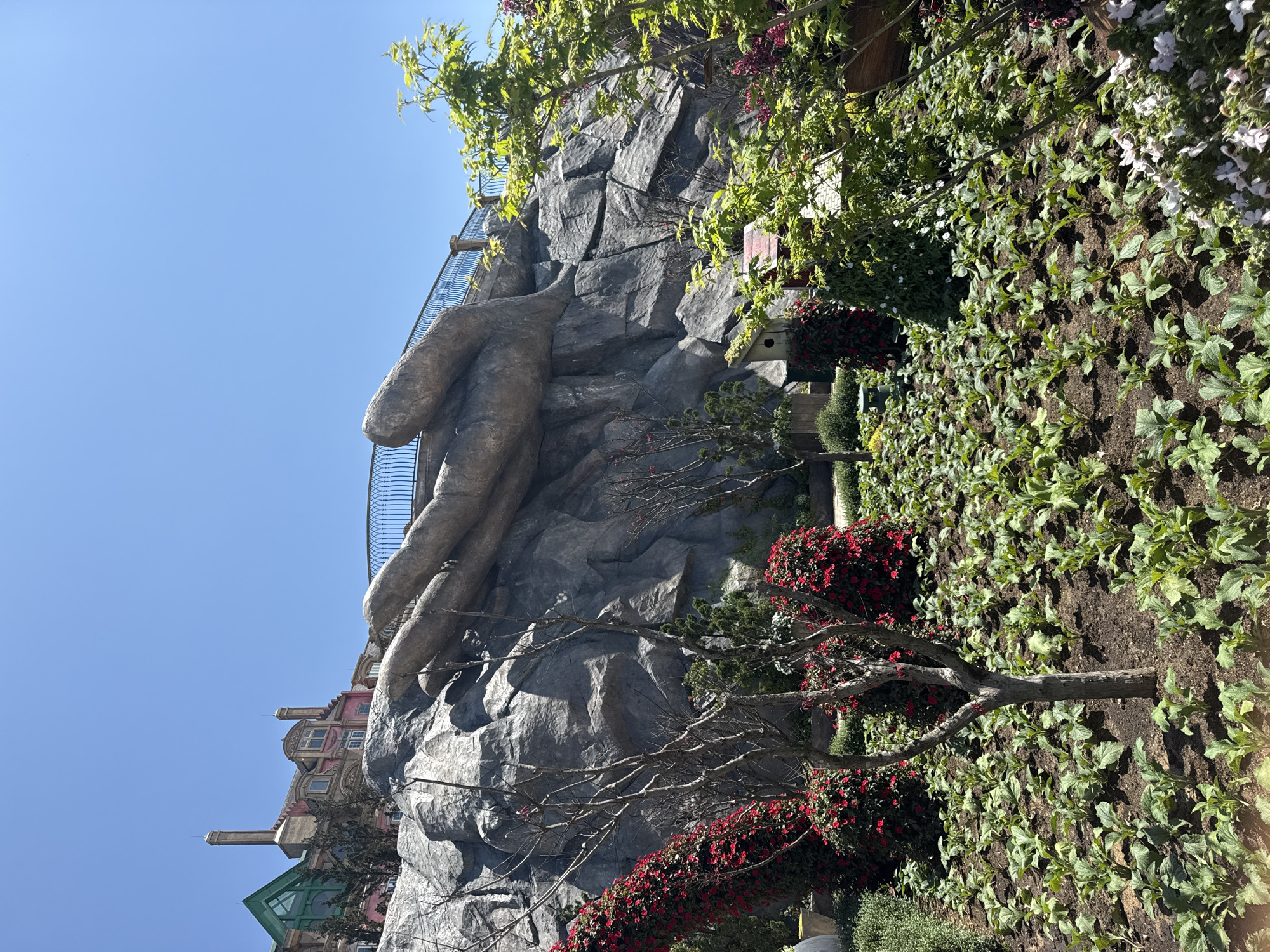 The image depicts a large, artificial rock structure with a prominent hand reaching out. The hand appears to be made of a grey, textured material and is positioned against a clear blue sky. At the base of the rock structure, there is a garden with a variety of plants and flowers, including red begonias. The garden appears to be well-maintained. In the background, sections of colorful buildings are visible, suggesting a cityscape. There is no indication that the image is taken in Đà Nẵng, Vietnam. No people are visible in the image.
