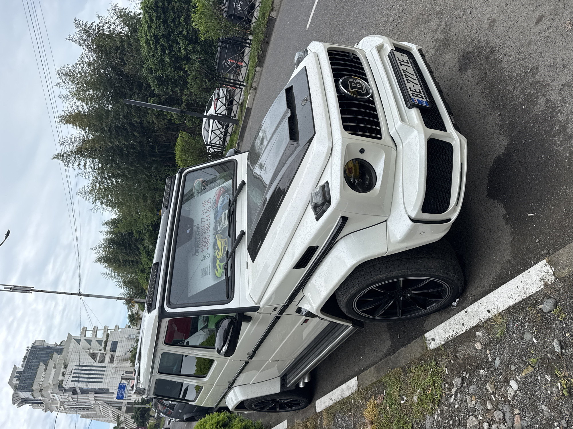The image depicts a white Brabus G-Class parked on the street. The car is parked along a roadside curb marked with a white line. The car's license plate reads "BE-777-TE". The vehicle is parked in Batumi, Georgia; a building characteristic of Batumi's architectural style is visible in the background. Trees and street foliage are visible behind the car. A sign is visible in the window of the car. The sign has phone numbers on it.
