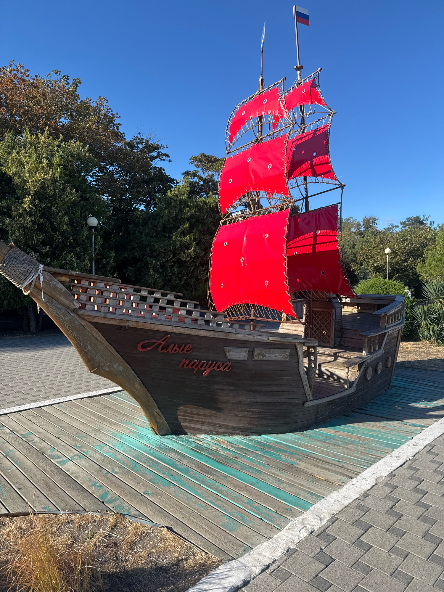 The image shows a wooden replica of a sailing ship with bright red sails, positioned on a wooden platform on what appears to be a sunny day. The ship is the primary subject, decorated with red fabric sails. The name "Алые паруса" (Alye parusa), meaning "Scarlet Sails" in Russian, is written in script on the side of the ship. In the background, there are trees and a clear blue sky. The ship is located in Anapa, Russia, as indicated by the text on the ship. The surrounding environment includes a paved walkway with interlocking grey pavers and some dry vegetation. The ship is an art installation or a decorative element, likely a landmark or attraction in the city. There are no people visible in the image. The overall scene suggests an outdoor public space.