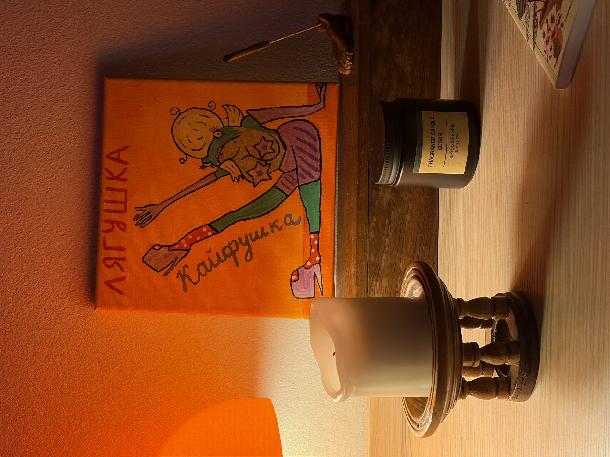 The image displays an indoor scene, likely a shelf or mantelpiece, illuminated by warm, artificial light, suggesting evening or nighttime.
The central subject is a framed canvas painting featuring a vibrant, cartoon-style depiction of a woman. She has blonde, curly hair and is shown in a dynamic, stretching or bending pose, looking over her shoulder. She wears a green top, striped purple bottoms, green tights, and pink polka-dotted socks paired with distinctive high-heeled platform shoes. Her chest is adorned with two star-shaped motifs. The painting’s background is a solid orange. Text written in Cyrillic is visible on the canvas: "ЛЯГУШКА" (Lyagushka), meaning "frog" or "froggy," and "Кайфушка" (Kaifushka), which could imply enjoyment or pleasure.
To the right of the painting, on a light-colored wooden surface, rests a dark-colored candle in a jar with a silver lid. Its yellow label reads "FRAGRANCE CANDLE" and "CEDAR," with Japanese text translating to "Fragrance Candle" and "Red Cedar." Further right, a white, unlit pillar candle is held within an ornate, dark wooden holder. A partially visible book, open to an illustrated or textual page, is also on the wooden surface.
Above the painting, a small, decorative wooden hook with a thin stick-like object, possibly a candle snuffer or tool, is attached to a darker wall. The wall to the left of the painting has a textured, warm orange surface, which catches the light. The overall ambiance is cozy and personal, with the various decorative items contributing to a relaxed setting. The scene is located in Chang Phueak, Thailand.