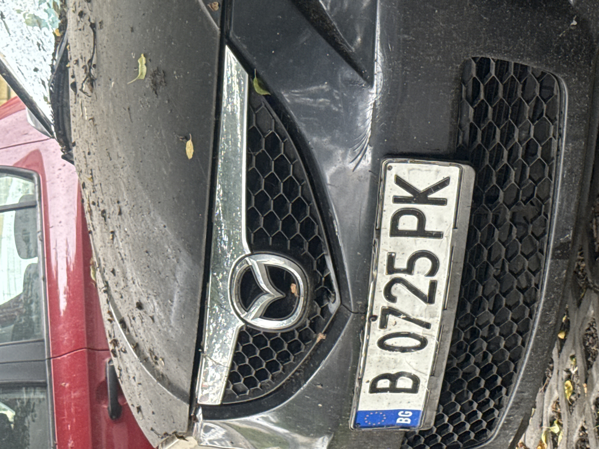 The image displays the front of a dark-colored Mazda car, heavily covered in dirt, dust, and scattered small leaves. The car's emblem is centrally visible on its honeycomb-patterned grille. To the right of the grille, a Bulgarian license plate is attached, clearly showing "B 0725 PK". The blue strip on the license plate features the European Union stars and the country code "BG", confirming its origin. A portion of a red vehicle is visible to the left of the Mazda. The scene is outdoors, likely during daytime, with natural light illuminating the dirty surface of the car. The car appears to be stationary and parked. The 'B' prefix on the license plate, along with the provided context, specifies the location as Varna, Bulgaria.