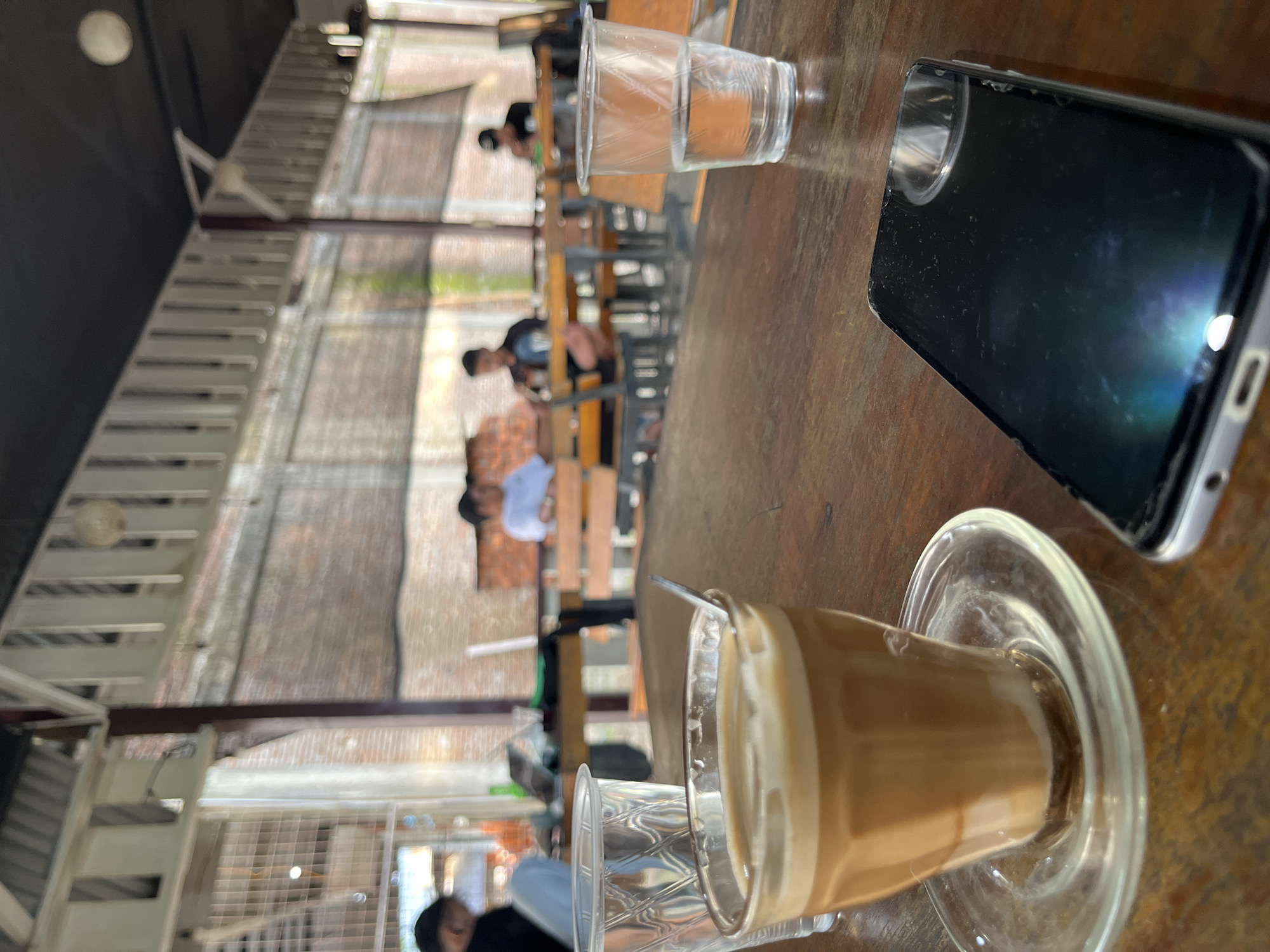 In the foreground of the image, a wooden table holds a glass of coffee with foam, a small glass saucer, a spoon, and a smartphone. To the left, another clear plastic cup with a textured pattern contains water. In the background, several people are seated at tables in what appears to be an outdoor cafe with a covered roof. The cafe has a rustic aesthetic with exposed brick walls and wooden benches. The lighting suggests it is daytime.