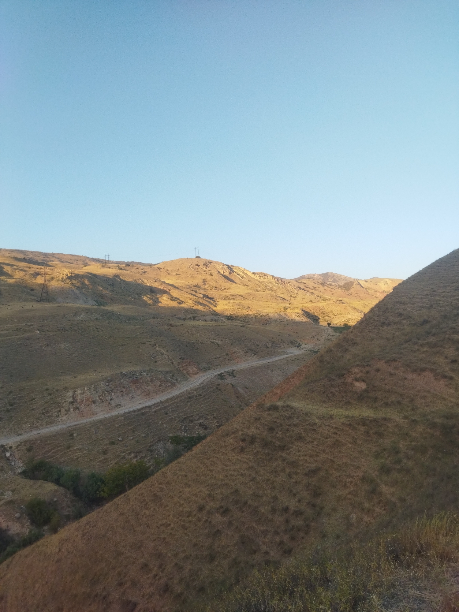 The image depicts a vast, rugged, and arid landscape under a clear, pale blue sky. The terrain is characterized by rolling hills covered in dry, golden-brown grass and sparse vegetation. A dirt road winds its way through the landscape, appearing to be the primary access route. In the background, a range of mountains stretches across the horizon, their slopes illuminated by the warm, low light of either sunrise or sunset, casting long shadows. Several electricity pylons are visible on the hilltops, suggesting the presence of infrastructure. The foreground is dominated by a steeply sloped hill also covered in dry grass. The environment appears remote and undeveloped, consistent with a rural or mountainous region. The location is identified as Mailuu-Suu City, Kyrgyzstan.
