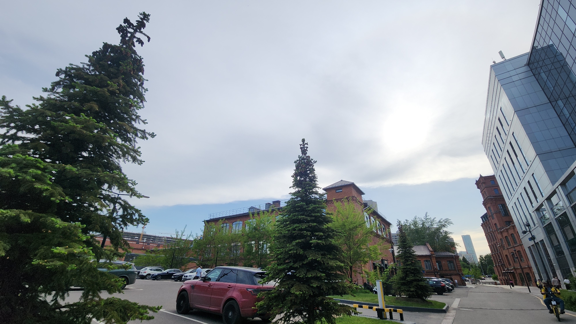 The image depicts a street scene in Moscow, Russia, featuring a mix of architectural styles. On the left, a large, mature evergreen tree dominates the foreground, adjacent to a parking lot with several cars, including a maroon Range Rover. In the background, a multi-story red brick building with green foliage is visible, suggesting a historical or renovated industrial structure. To the right, a modern glass and steel building rises, contrasting with the brick structures. Further down the street, more red brick buildings are seen, creating a visual blend of old and new architecture. A person on a scooter rides down the street on the right side of the image. The sky is partly cloudy, indicating a fair weather day.