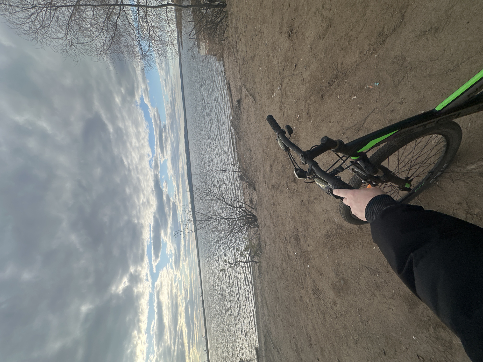 The image shows a bicycle on a dirt path beside a body of water, likely a river or lake. The handlebars of the black and green bicycle are visible, with a hand holding the grip. The background includes water reflecting a cloudy sky. The location is Chaikovsky, Russia. The photograph appears to be taken from a first-person perspective.