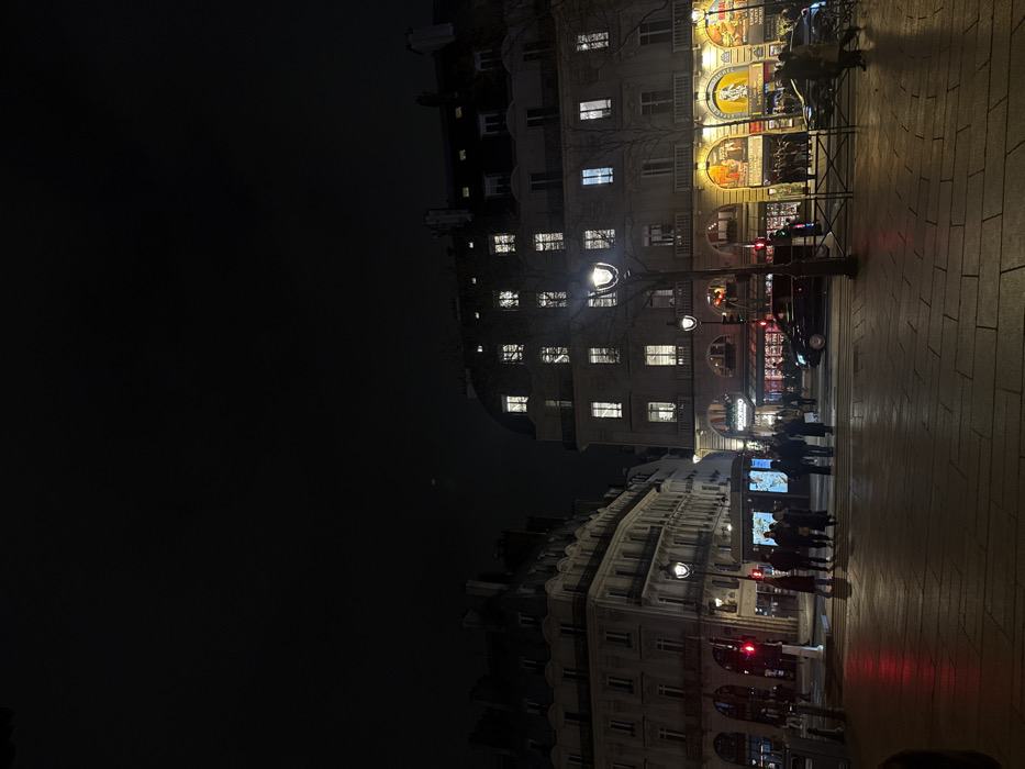 The scene depicts a bustling urban street at night, specifically at an intersection on one of the Grand Boulevards in Paris, France, likely near Boulevard Montmartre. The sky is dark, contrasting with the numerous artificial light sources.
In the mid-ground, a group of approximately 10-15 individuals, predominantly adults, are congregated on a paved pedestrian crossing, some appearing to wait. To their left, a traffic light displays red. Another illuminated glass kiosk or bus stop shelter is visible nearby. Further to the right, a dark sedan, possibly a taxi, is present in the street.
The background features two multi-story Haussmannian buildings. The building on the left has several dimly lit windows and balconies on its upper floors. The building on the right is more brightly illuminated, with numerous windows showing interior light. Its ground floor houses commercial establishments, including one with prominent illuminated posters, one displaying "ROXANE" and another referencing "GRAND BOULEVARD", suggesting a theater or entertainment venue.
Several ornate Parisian street lamps illuminate the area, with one prominent lamp in the mid-ground next to a bare-branched tree. The foreground consists of a wide, light-colored paved area, reflecting some of the ambient light. Metal barriers are visible on the right side of the street. The overall environment is active, indicating a vibrant city night scene.