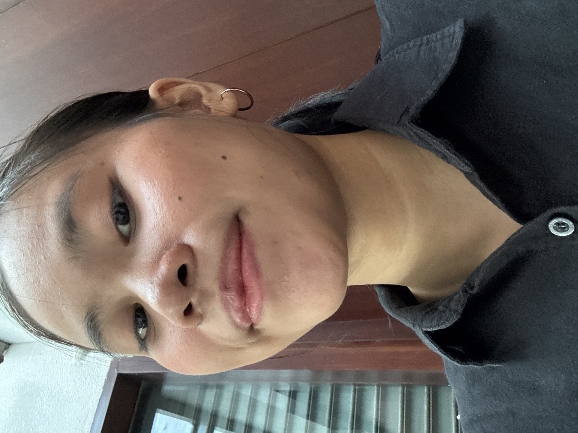 A medium shot, close-up selfie of a young woman is captured from the shoulders up. She is wearing a black collared shirt, unbuttoned at the top. A small, gold hoop earring is visible in her right ear. Her dark hair is pulled back. The background consists of a light-colored wall on the left and a wooden surface on the right. The image appears to have been taken indoors. The woman's face is turned slightly towards the camera, with her lips partially open and a faint smile. Her eyes are looking directly at the camera. The lighting is natural, casting soft shadows. The overall scene is casual and personal. There is no discernible location in South Jakarta, Indonesia.