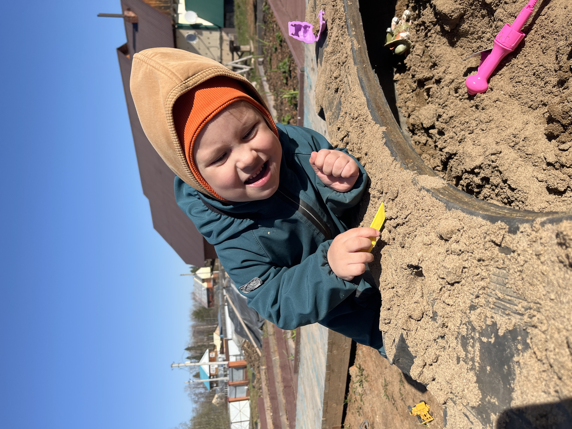 A young child wearing a tan hooded jacket with an orange hat is playing in a sandbox made from a large black tire filled with sand. The child is smiling with visible teeth and holding a yellow plastic toy. Other plastic toys, including a pink shovel and a purple mold, are also in the sandbox. The sandbox is positioned outdoors, likely in a backyard or garden area. In the background, a variety of buildings and trees are visible under a clear blue sky, indicative of a sunny day. The location is Chaikovsky, Russia.