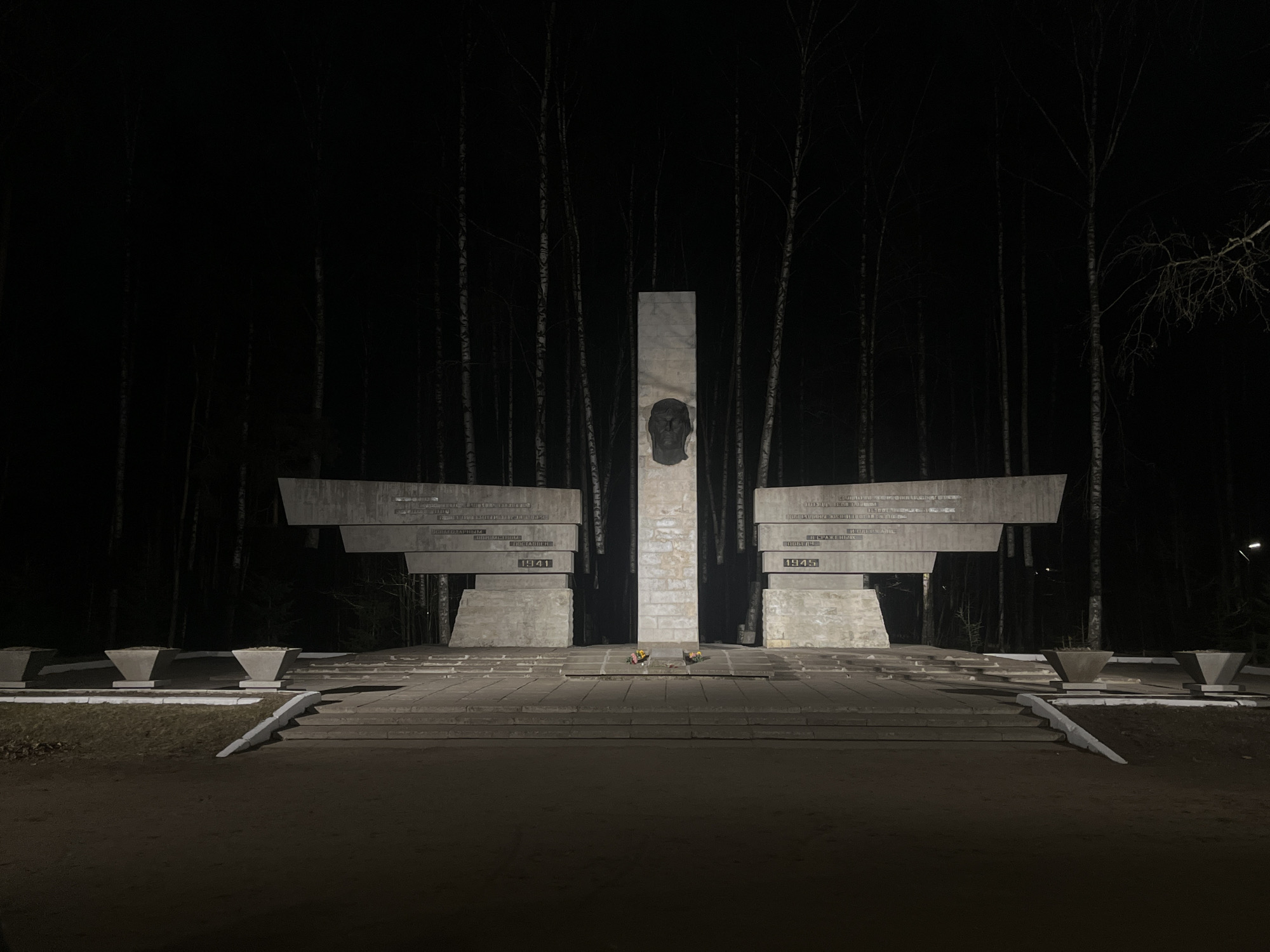 The image depicts a war memorial at night, likely in a park or forested area. The memorial features a central pillar with a dark bas-relief of a human head. On either side of the pillar are concrete structures with inscriptions in Cyrillic and the dates 1941 and 1945, suggesting the memorial commemorates victims or events related to World War II. The base of the memorial is a raised platform with steps leading up to it, where flowers have been laid. In the foreground, there are empty flower pots, and the background consists of a dense forest. The overall impression is somber and commemorative, with the darkness emphasizing the gravity of the subject. The location is the memorial in the city of Saint Petersburg, Russia.