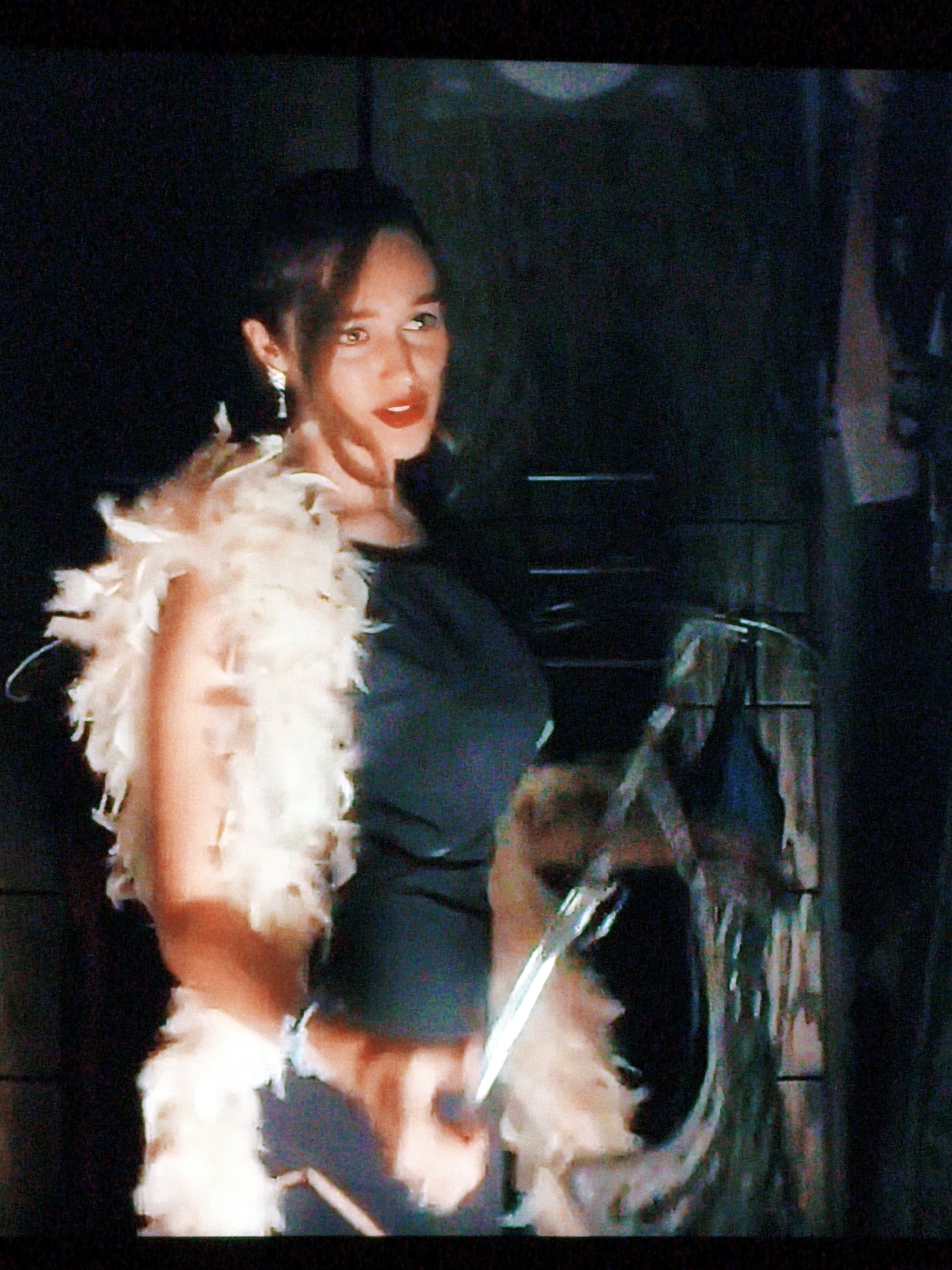 The image depicts a woman with dark hair, wearing a black dress and a white feather boa. She is adorned with earrings. Her makeup includes red lipstick. The background is dark and appears to be indoors, with some indistinct objects and vertical lines suggesting a wall or structure. There are no visible elements that indicate the location in Vladivostok, Russia.