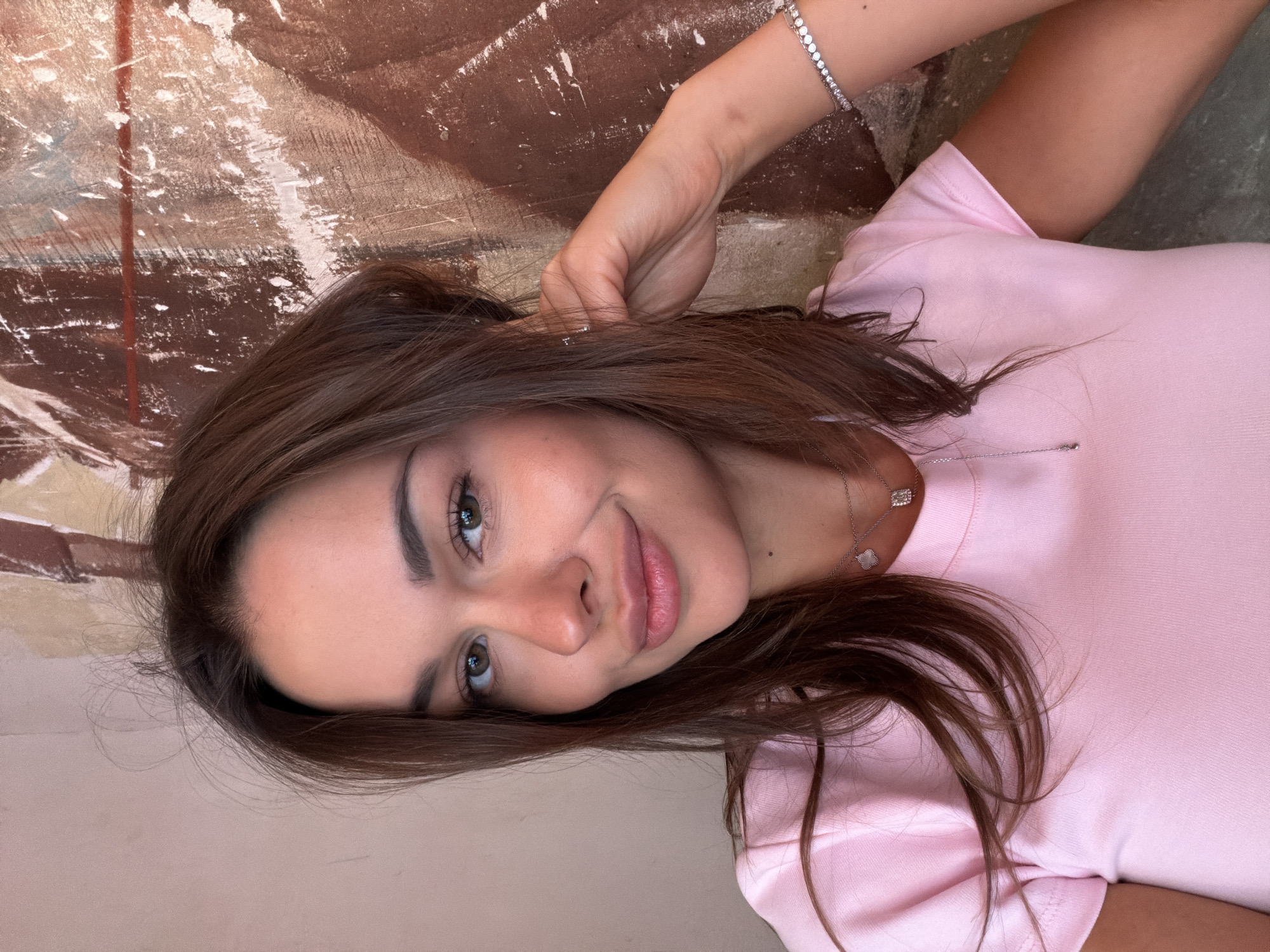 The image is a portrait of a young woman with long, brown hair and green eyes. She is wearing a light pink t-shirt and a silver necklace with two pendants. Her right hand is raised to her ear. She is smiling and looking at the camera. The background appears to be a textured wall with brown and cream colors. There is no indication of the location being Saint Petersburg, Russia.