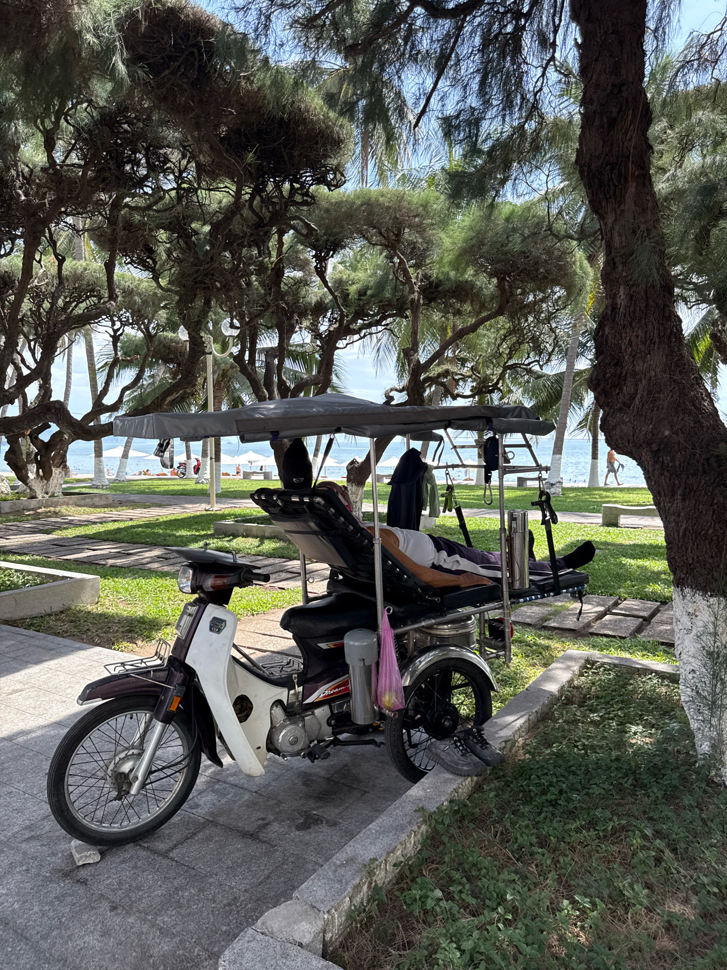 A modified motorcycle with a canopy and a reclining seat is parked on a paved pathway adjacent to a grassy area. A person is lying on the reclining seat, facing towards the left. The motorcycle has a dark frame and a white front fairing with some red and black decals. A pink plastic bag is hanging from the side of the motorcycle. In the background, there is a view of the sea, with several white umbrellas and people on the beach. Palm trees and other foliage are visible in the scene. The area appears to be a public space, likely a park or promenade along the coast of Nha Trang, Vietnam.